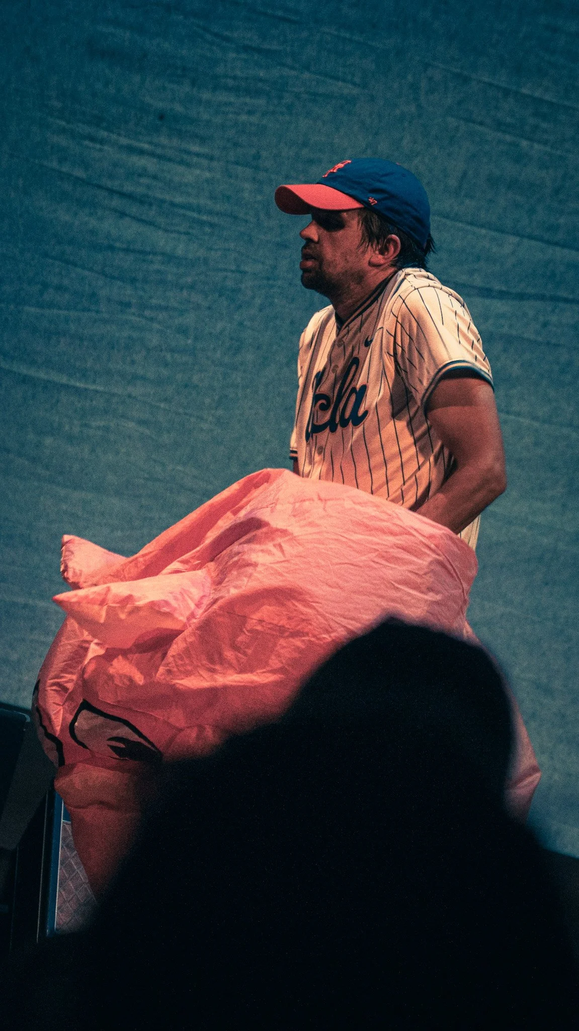 A man wearing a baseball cap and a baseball jersey holding a large orange bag, standing against a dark textured background.