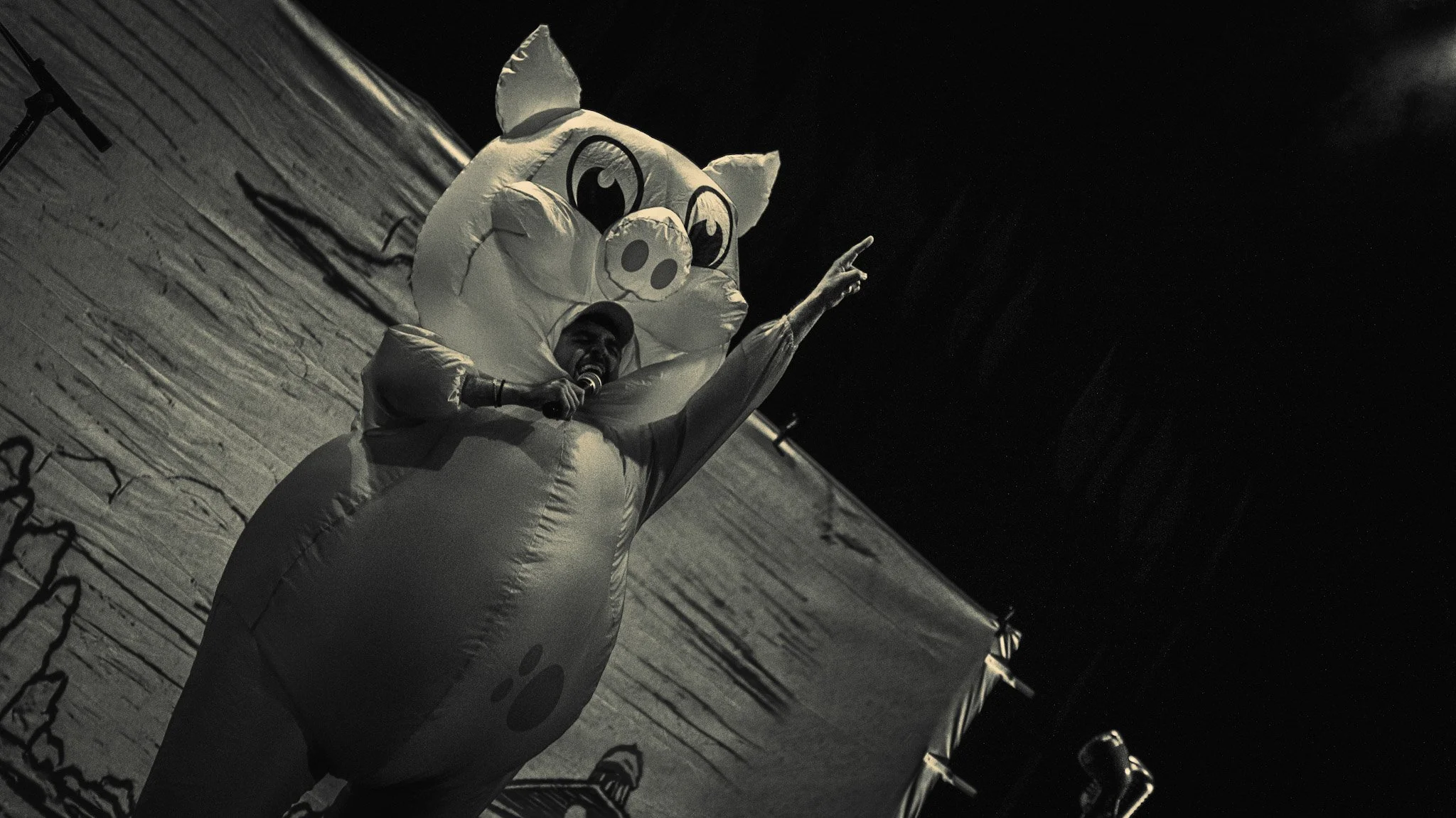 Person in an inflatable pig costume holding a microphone and pointing upward on a wooden stage at night.