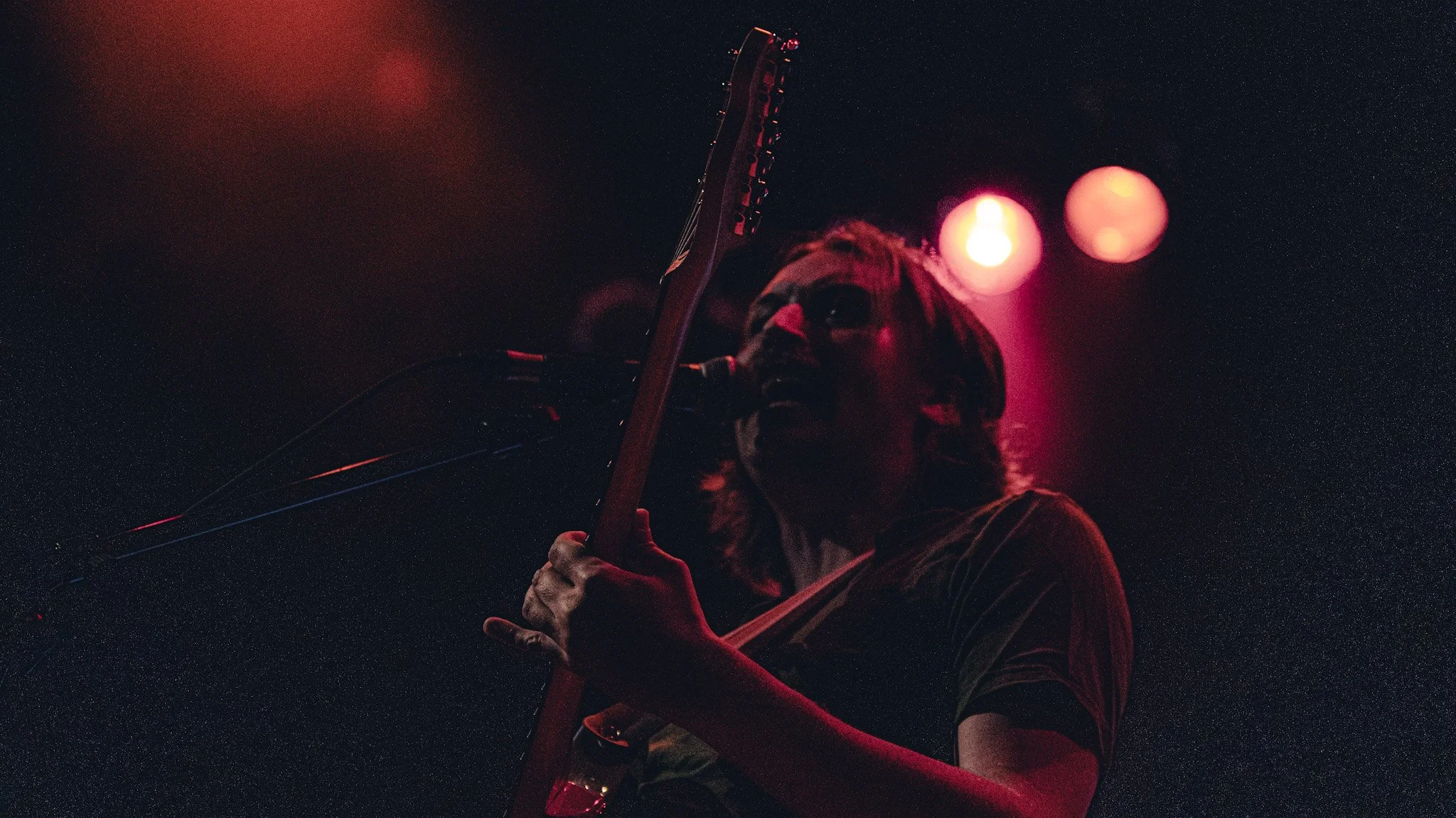 A person is playing an electric guitar on stage at night, illuminated by pink and red lights, with a starry sky in the background.