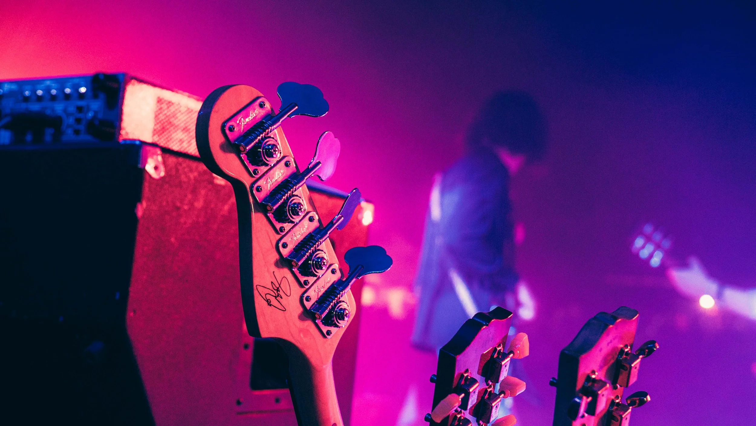 A close-up of a pink electric guitar headstock with tuning pegs, against a blurred background with purple and pink lighting.