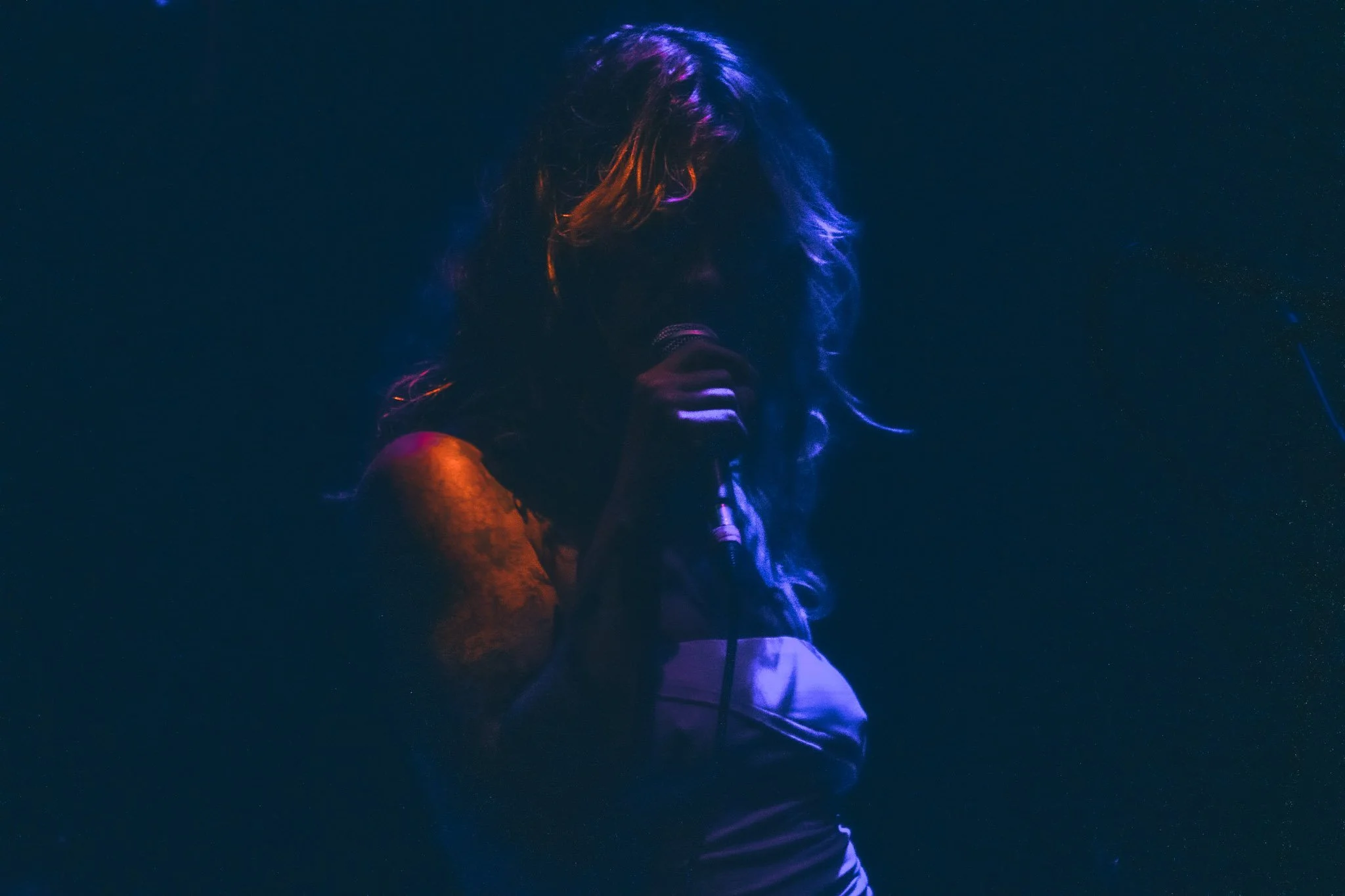 Person with long hair holding a microphone, illuminated by colorful stage lighting, on a dark background.