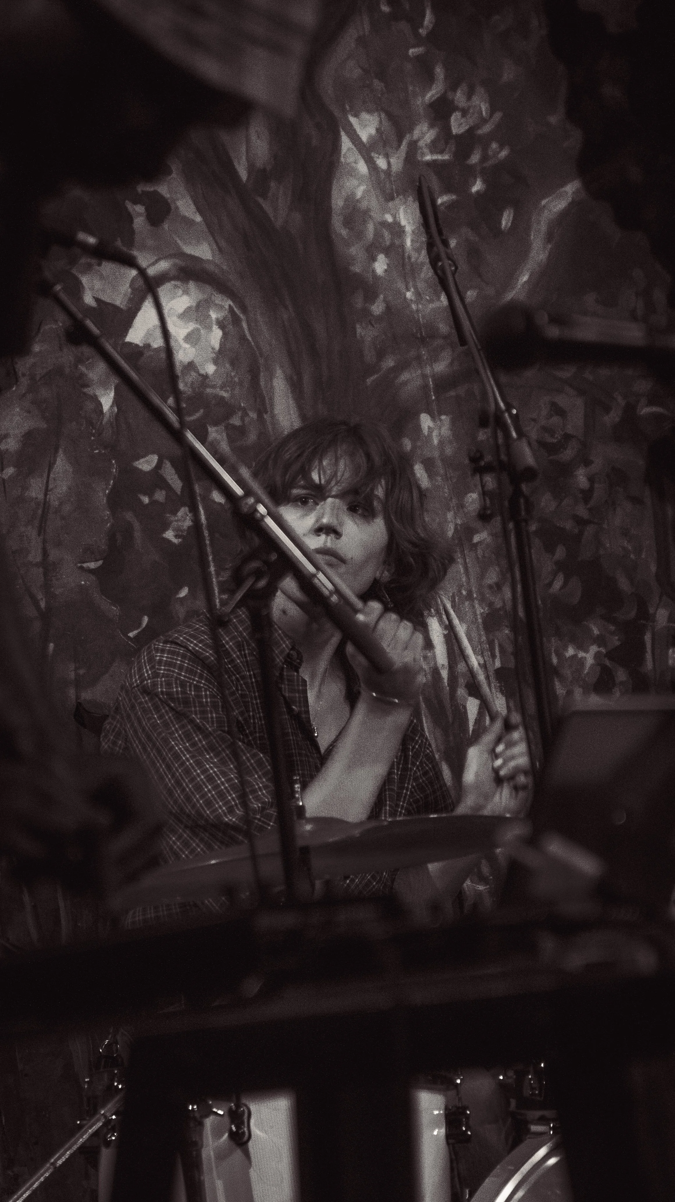 A woman with short hair and a checkered shirt sitting at a drum set, holding drumsticks, in a music studio with a leafy wallpaper background.