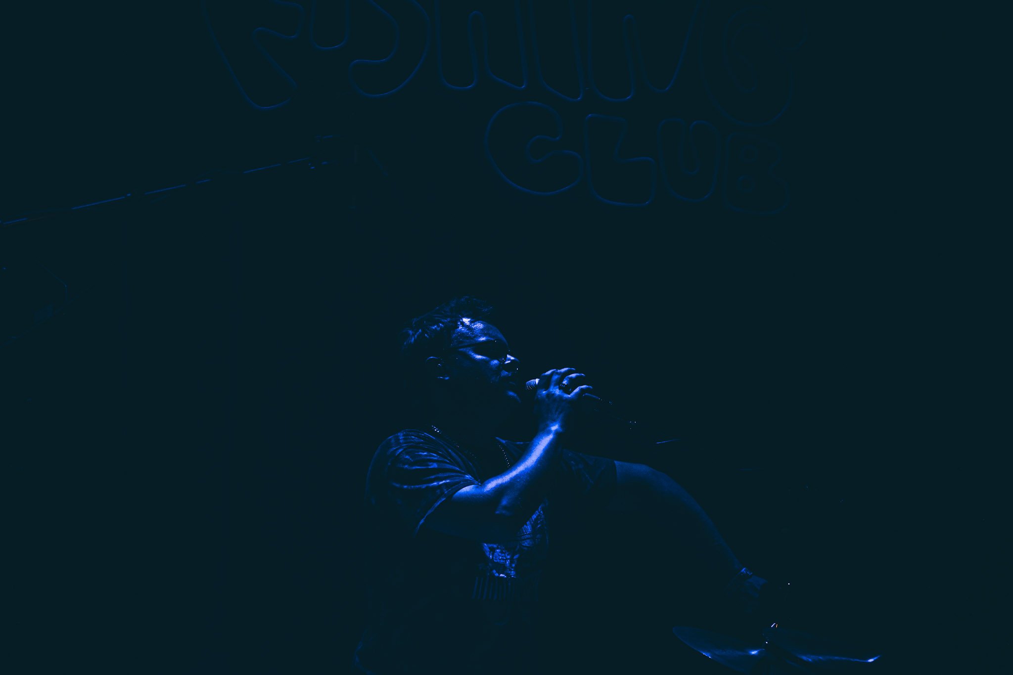 A man performing on stage under blue neon lights, with a neon sign above him