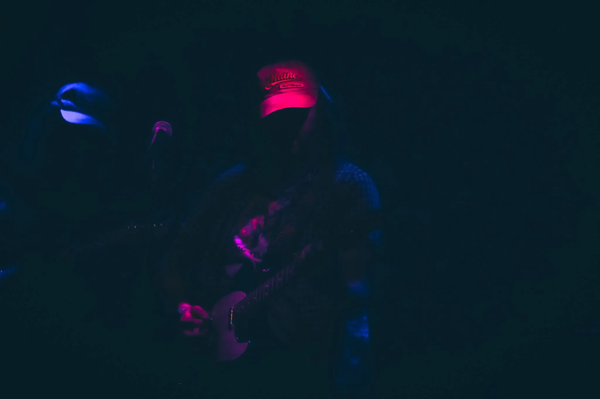 Dark image with faint pink and blue neon lights illuminating a person's face, wearing a baseball cap and looking downward.