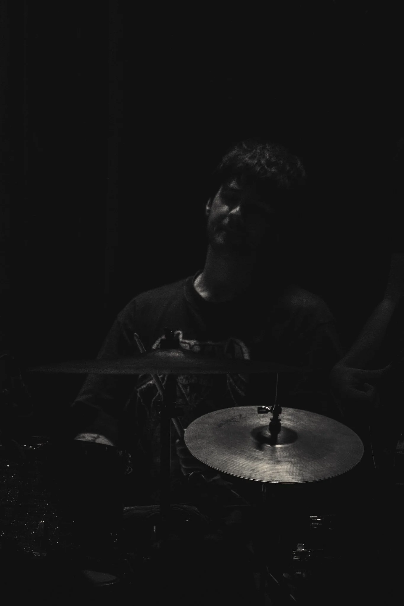 A person with curly hair playing drums in a dark room with minimal lighting.