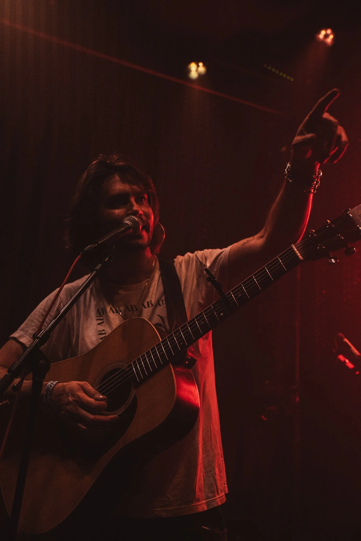 A person playing an acoustic guitar and singing into a microphone on a dimly lit stage with red lighting.