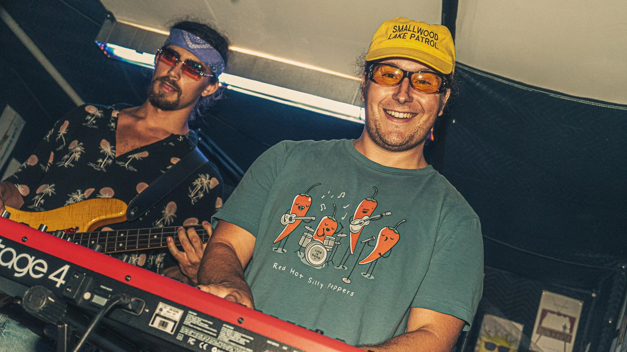 Two men are playing music. The man on the right wears an orange cap labeled 'Smallwood Lake Patrol', glasses, and a gray t-shirt with cartoon hot peppers playing instruments. The man on the left wears a blue headband, red sunglasses, a floral shirt, 