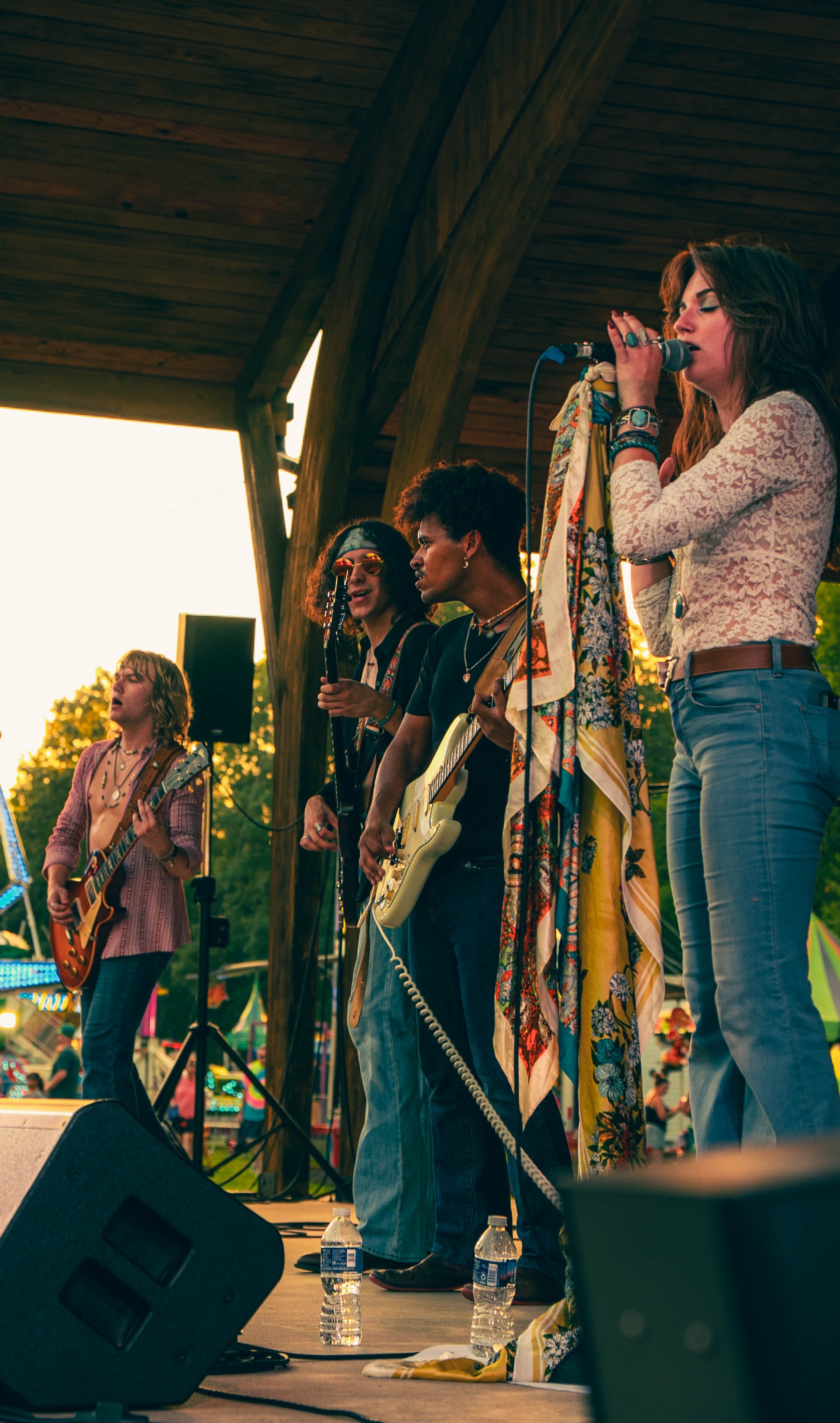 A band of five women performing on an outdoor stage during sunset, with three of them singing into microphones and playing guitars. They are dressed in casual, bohemian styles, surrounded by musical equipment and bottles of water, with a wooden pavil