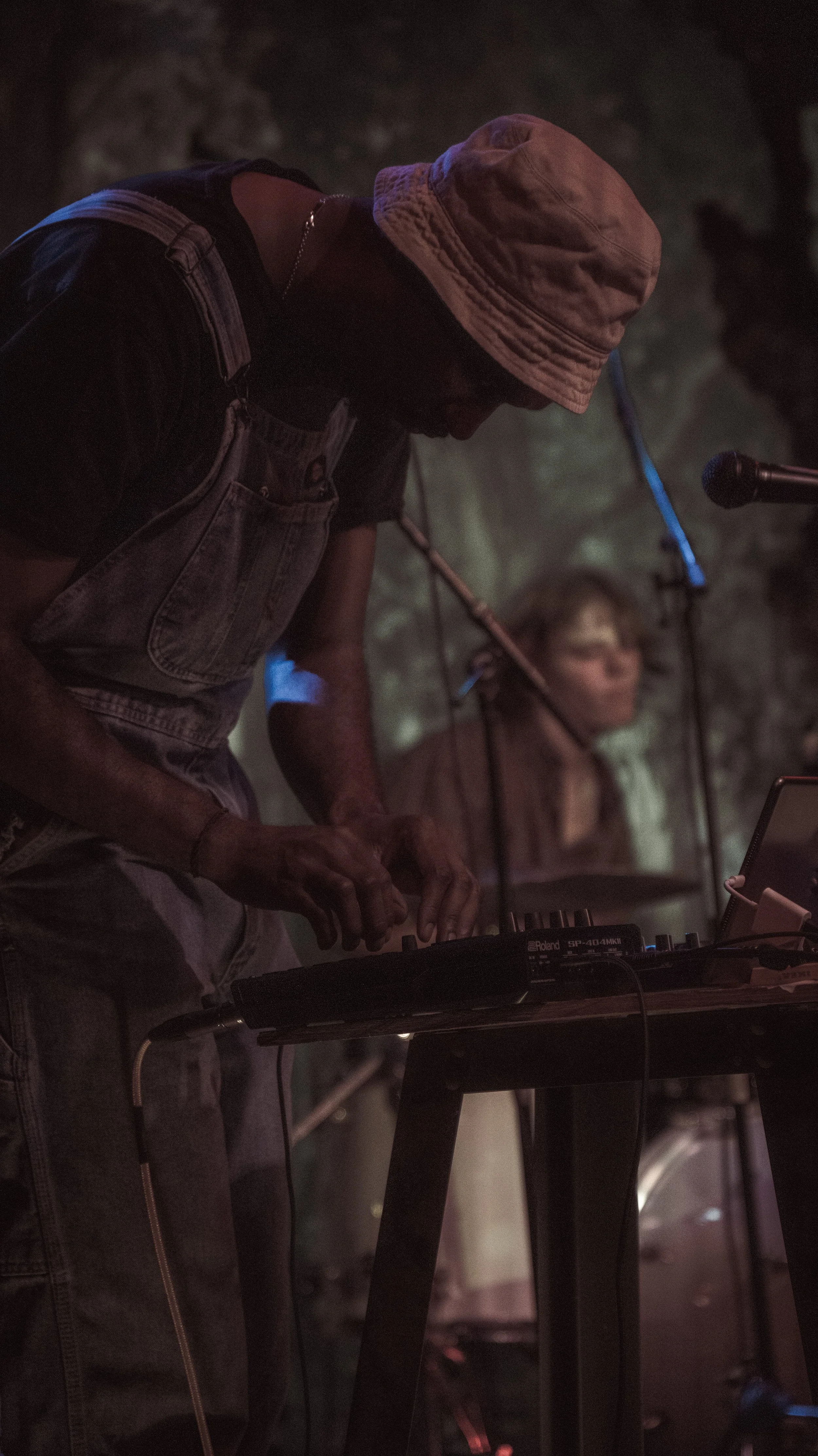 A person wearing a light-colored bucket hat and denim overalls, playing a keyboard at a dimly lit music performance.