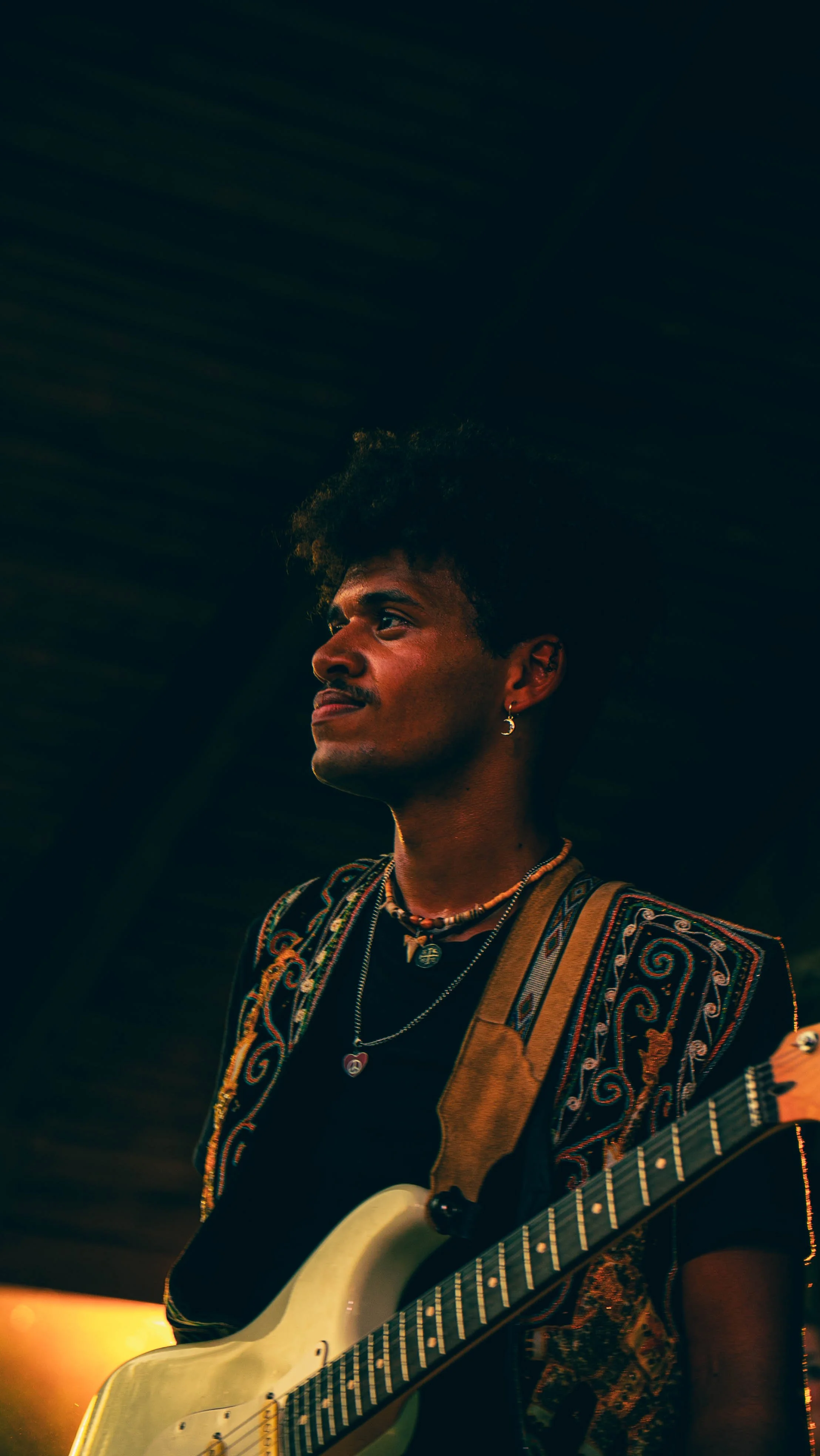 A young person with curly hair and earrings, wearing a colorful embroidered shirt, playing an electric guitar on stage with warm lighting.