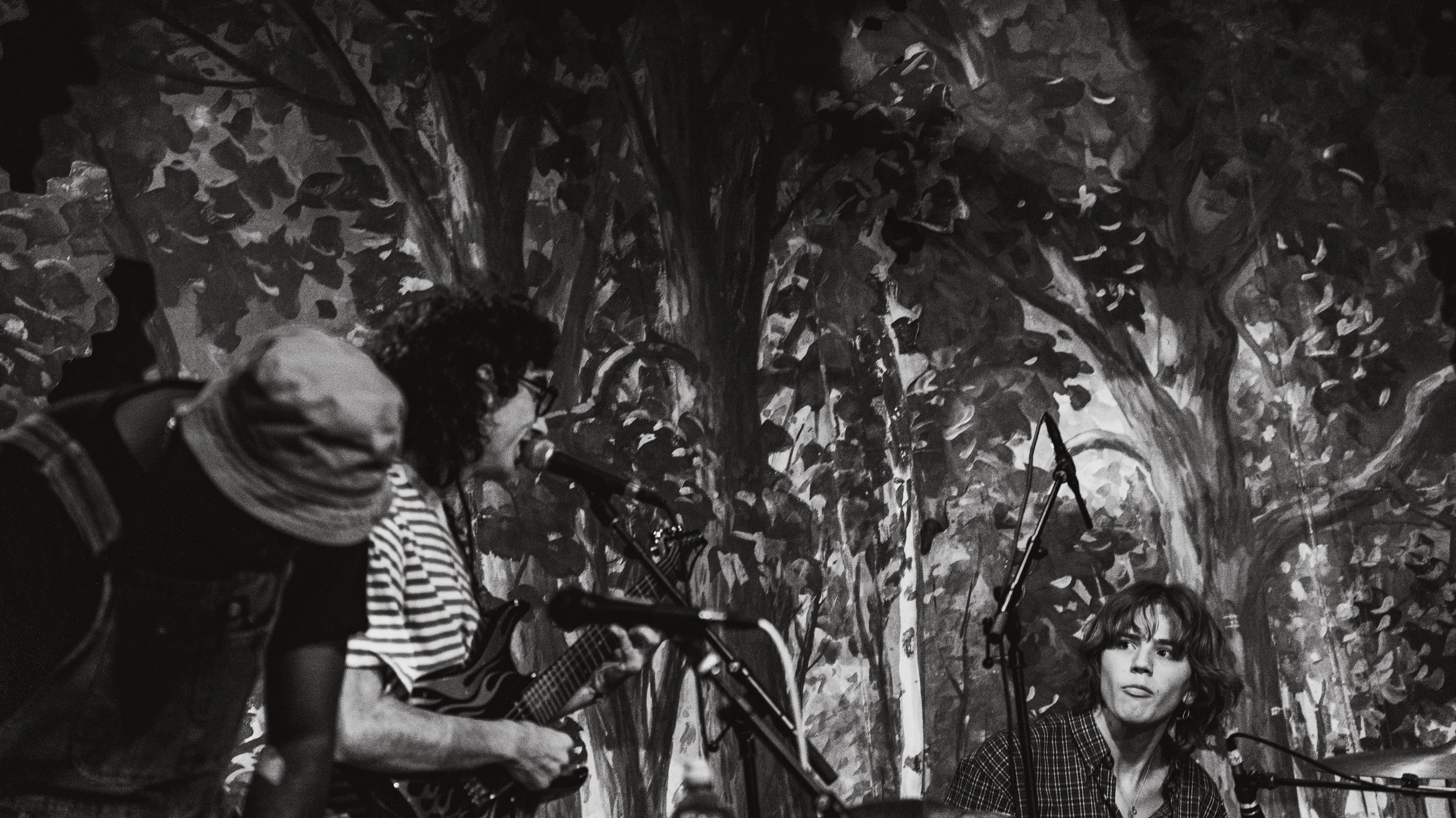 A black and white photo of two women performing music on stage with a painted backdrop of trees. One woman is holding a guitar and singing into a microphone, while the other woman is looking at her.
