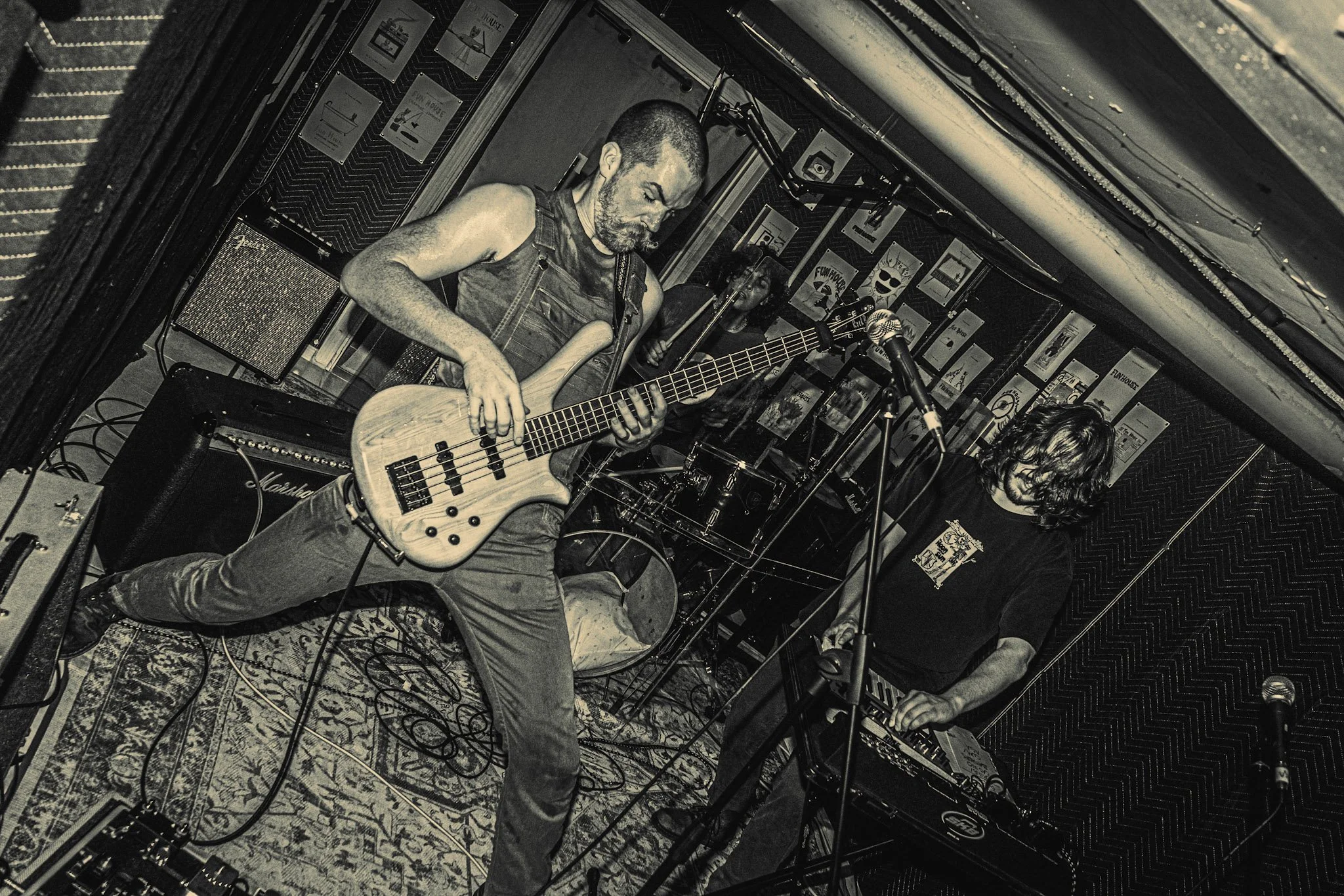 Musicians performing on a small stage, one playing a bass guitar and another singing into a microphone, in a venue decorated with posters.