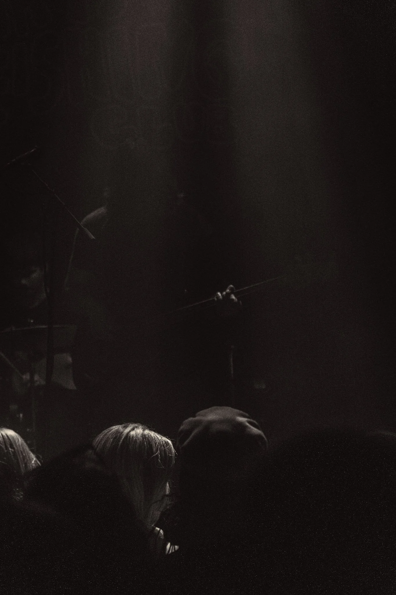 Dark scene with the outline of a crowd of people at the bottom, stage lighting and equipment visible at the top, and a person standing on stage in the distance.