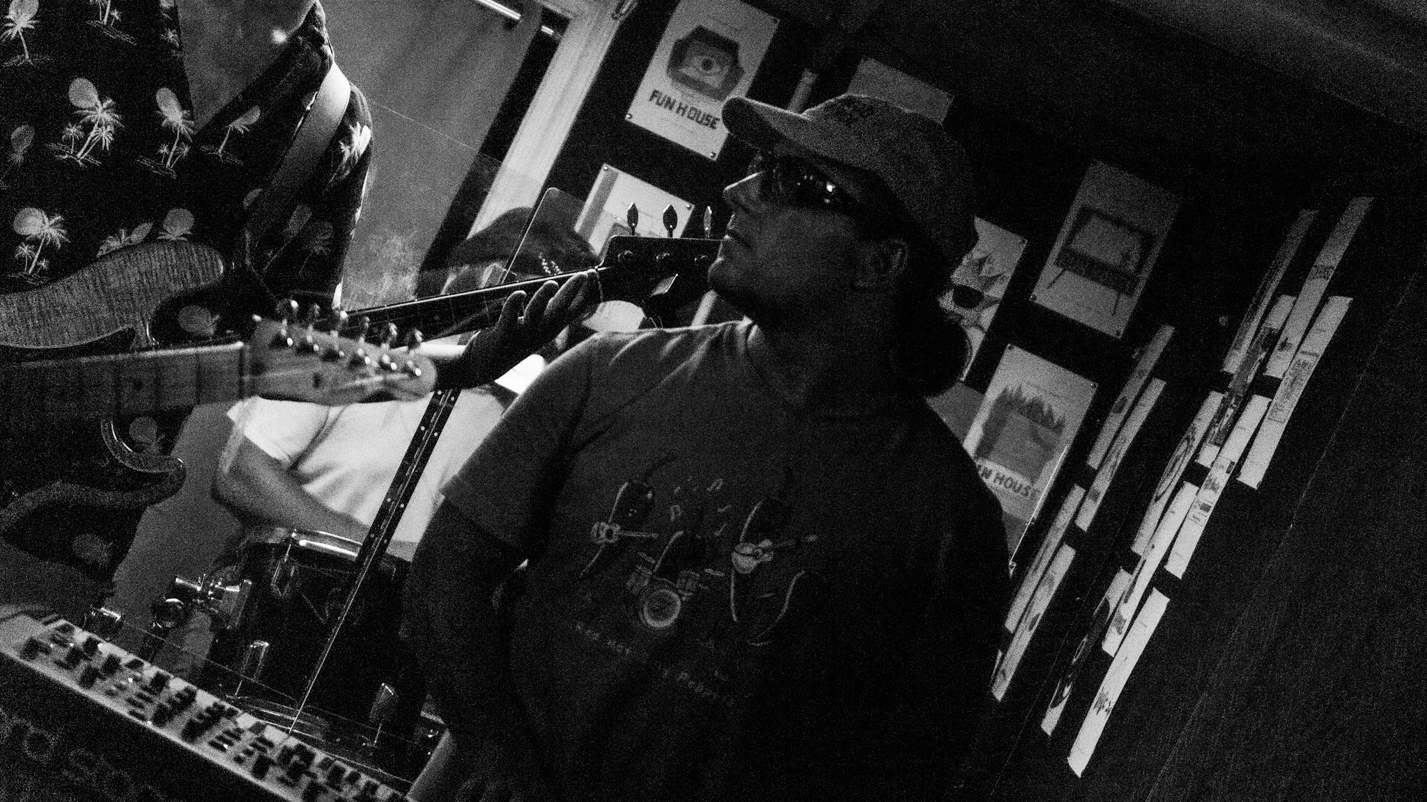 A person wearing sunglasses and a hat is playing an acoustic guitar in a room filled with posters and framed pictures on the walls. The image is in black and white.