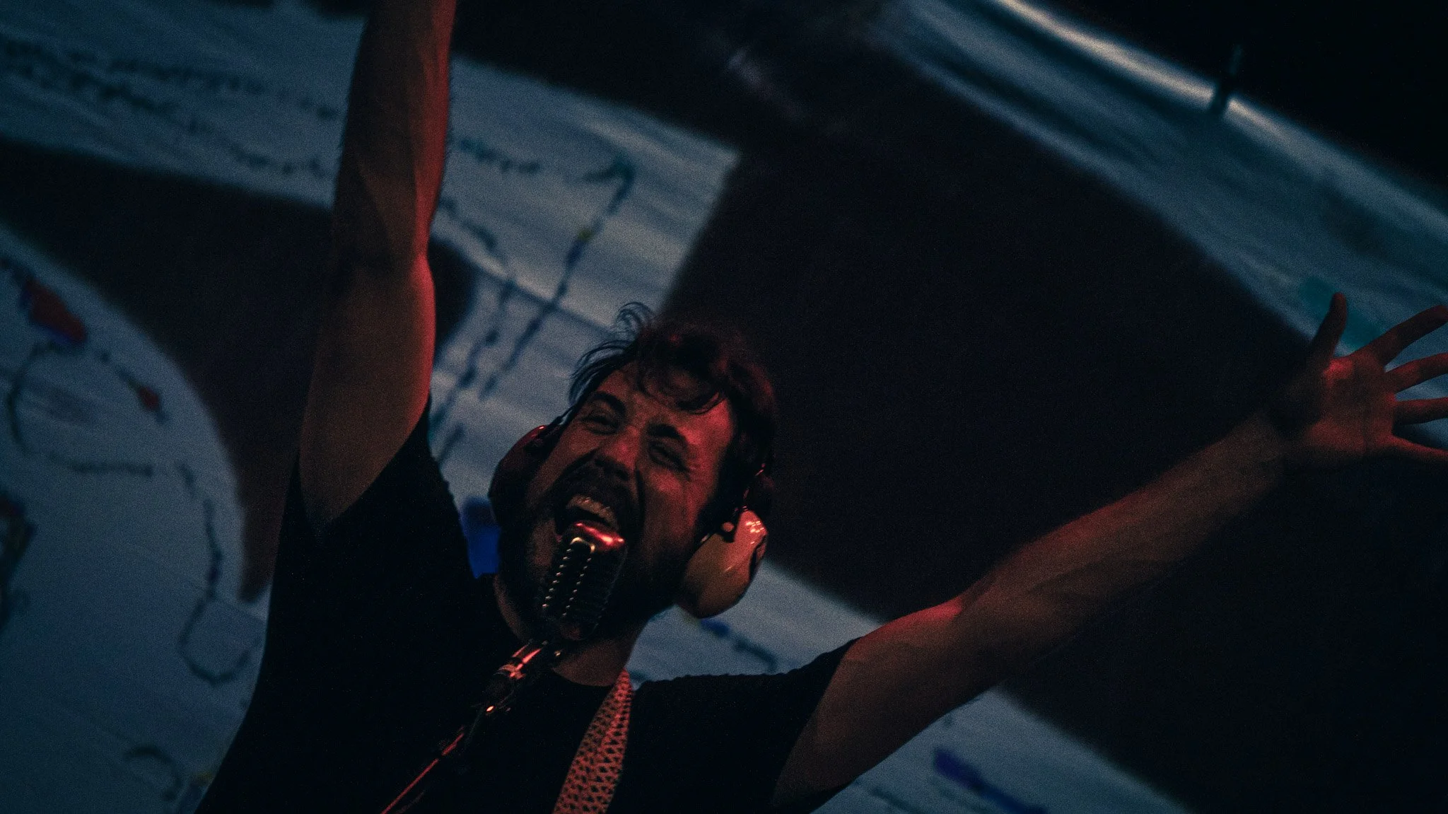 A person with short dark hair and a beard, wearing headphones and a headset with a microphone, is smiling and reaching upward with one hand. The scene appears to be in a dimly lit room with faint blue and red lighting.