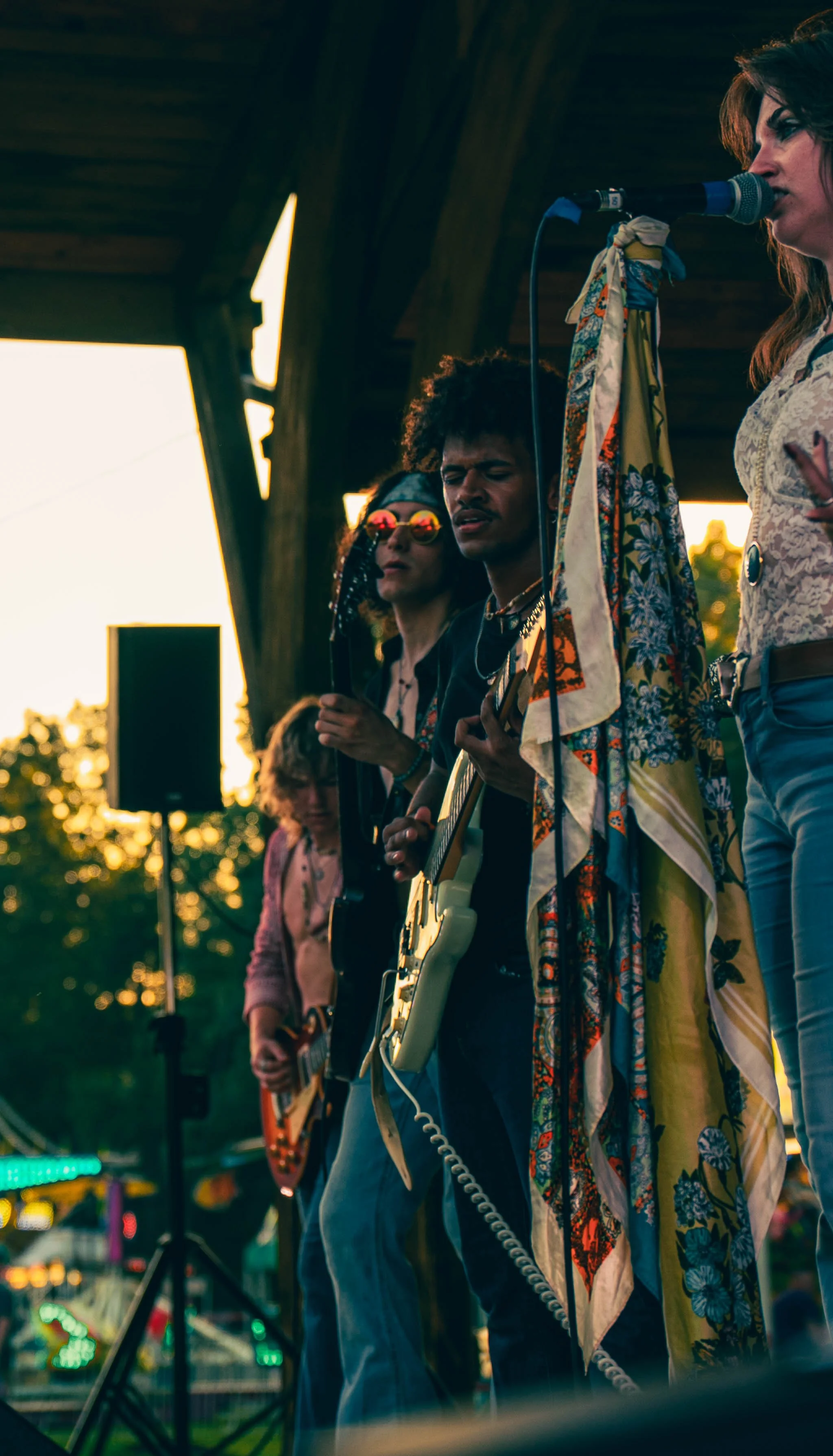 Musicians performing on stage at an outdoor event during sunset, including a woman singing into a microphone, a man playing guitar, and others in the background playing instruments.
