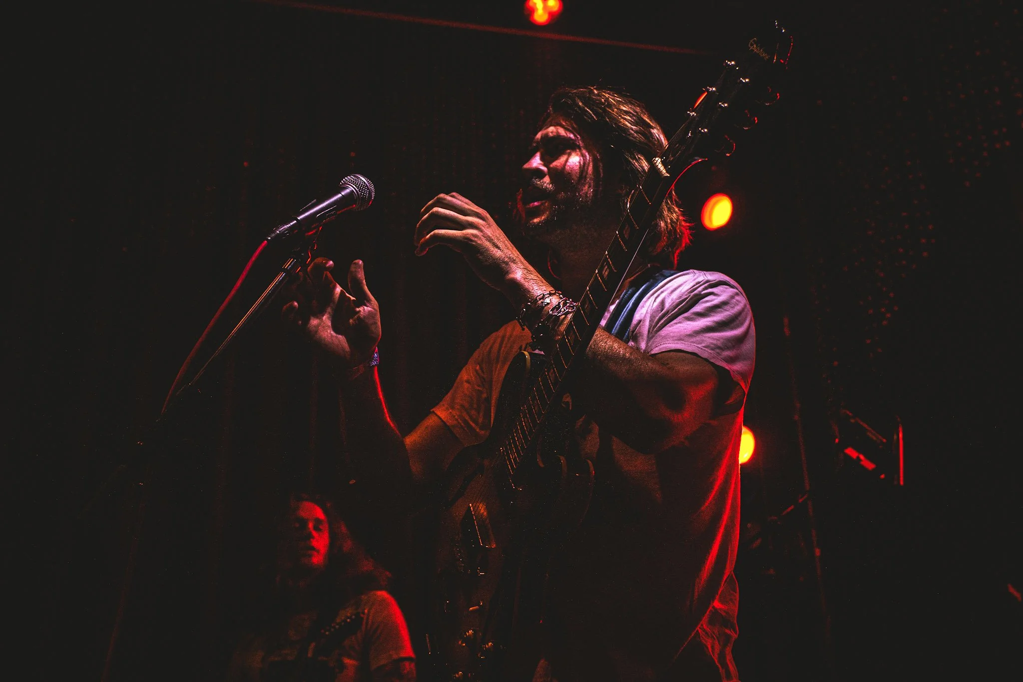 A person with long hair, sunglasses, and a white shirt playing an acoustic guitar and singing into a microphone on a dark stage with red lighting.