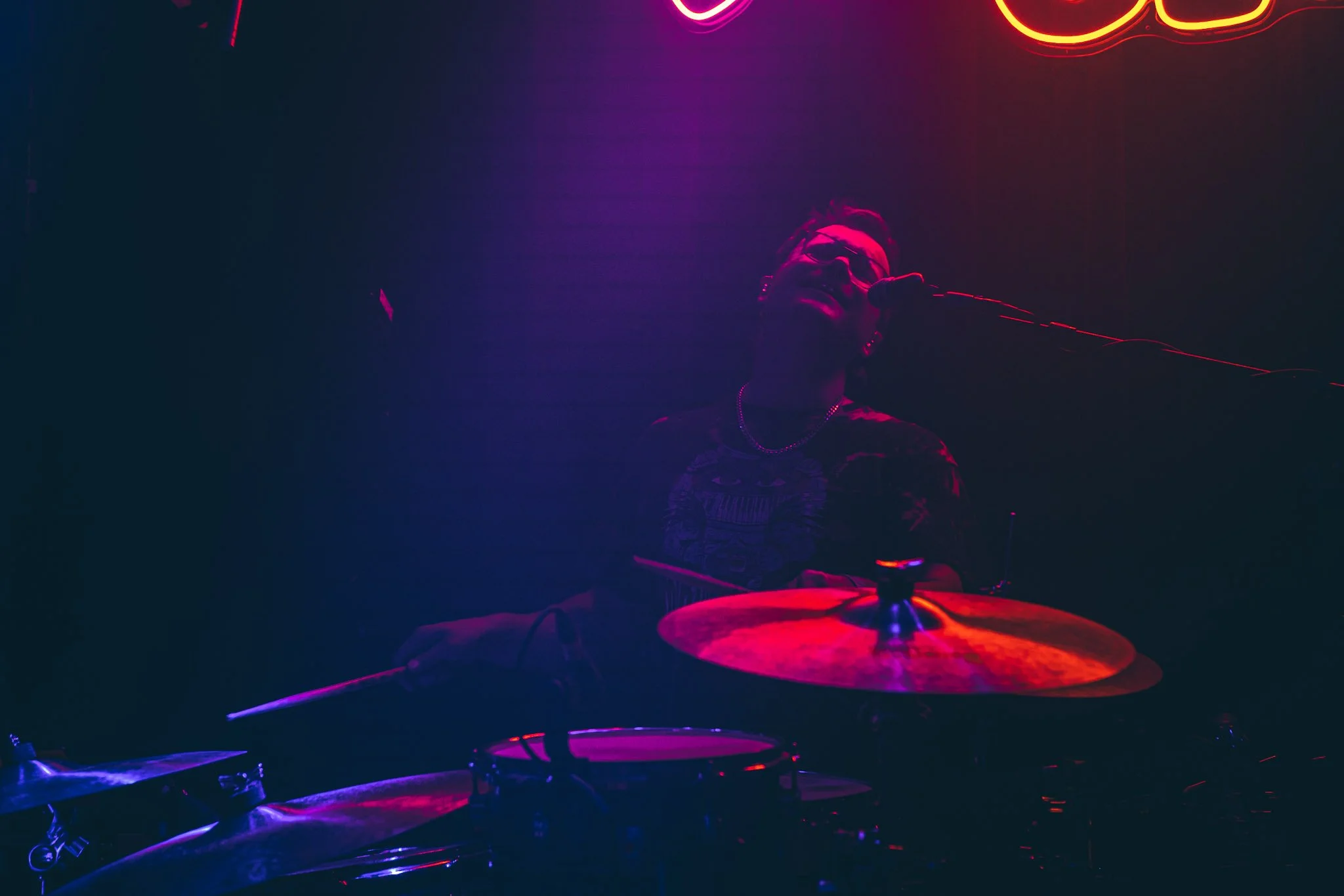 A person with glasses singing into a microphone in a dark room with neon pink and red lights, seated behind a drum set.