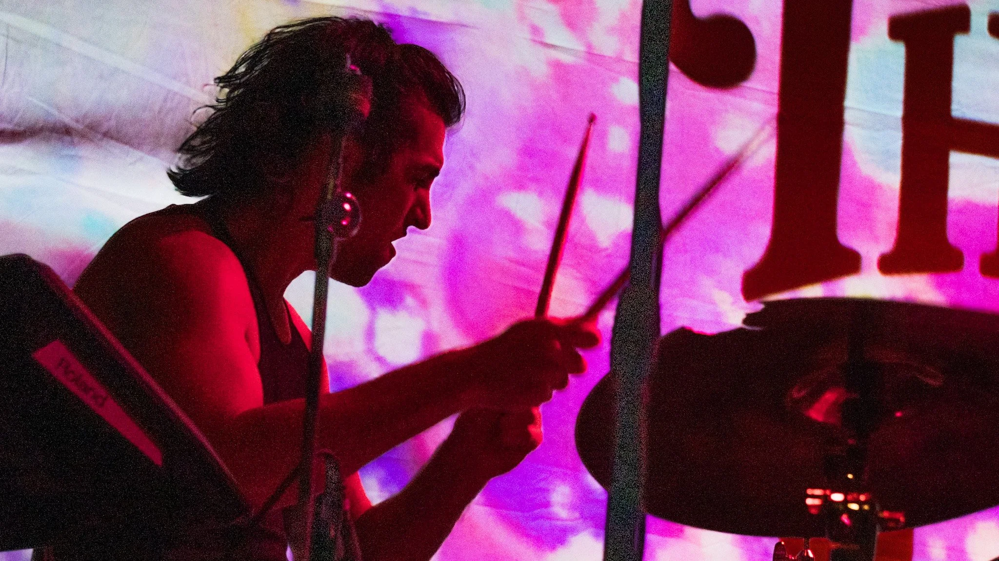 A woman playing drums at a concert, with a colorful, abstract background projecting purple, pink, and blue hues.