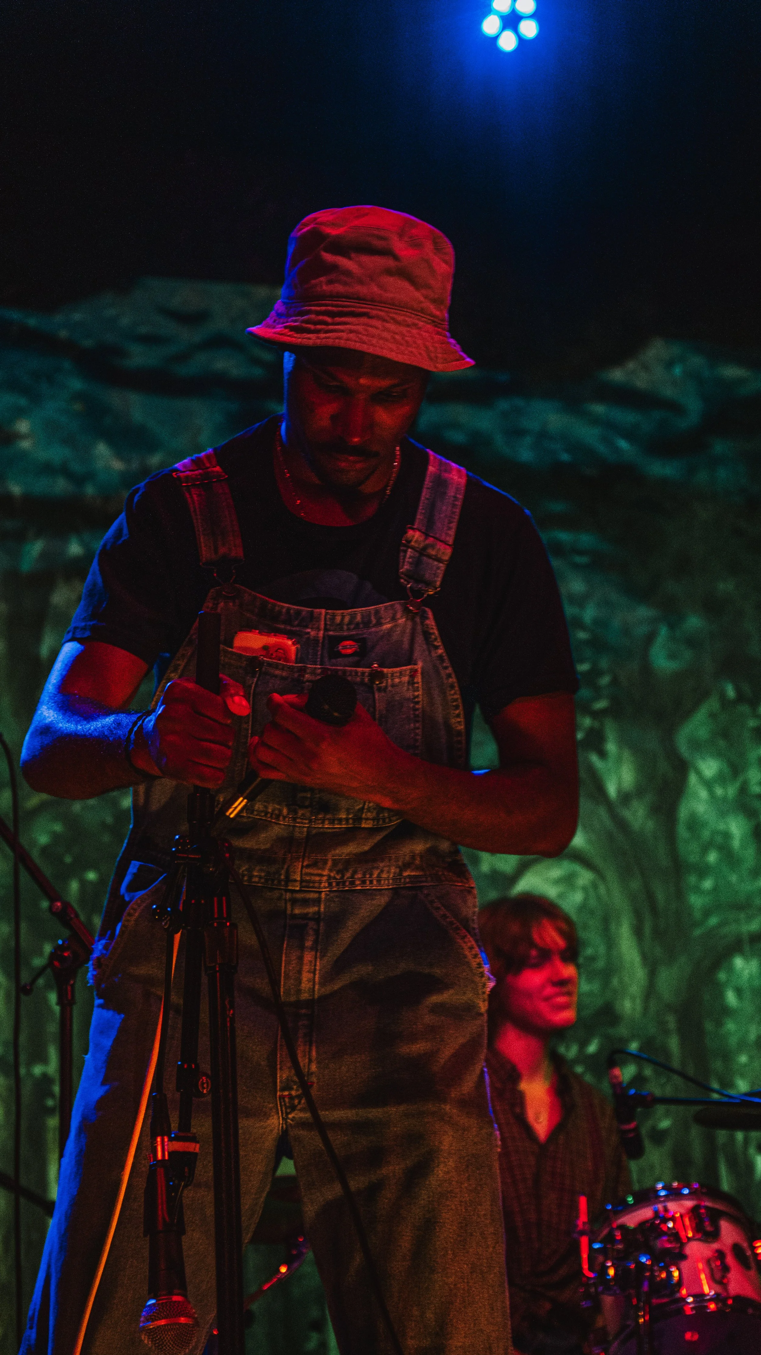 A person wearing a red bucket hat and denim overalls is on stage at a concert or event, adjusting a microphone. There are colorful stage lights and another person playing drums in the background.