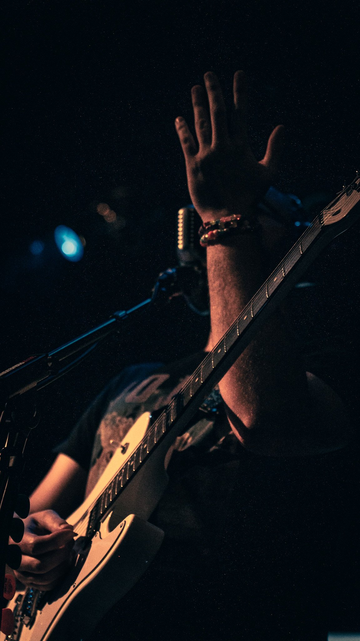 A person playing an electric guitar on a dark stage with a hand raised, wearing a bracelet and a wristwatch.