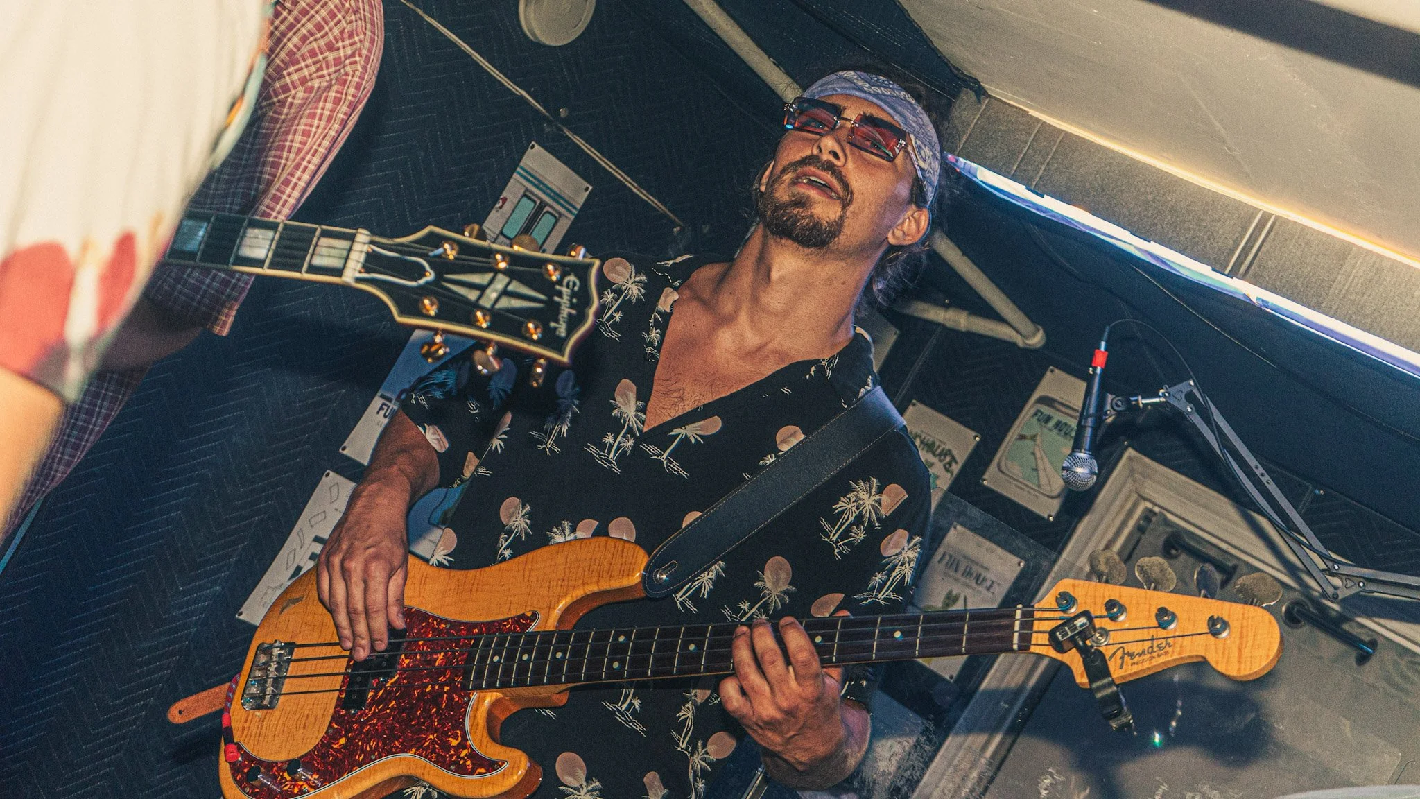 A man with sunglasses, a bandana, and a mustache playing an orange electric guitar in a room decorated with posters and a mural.