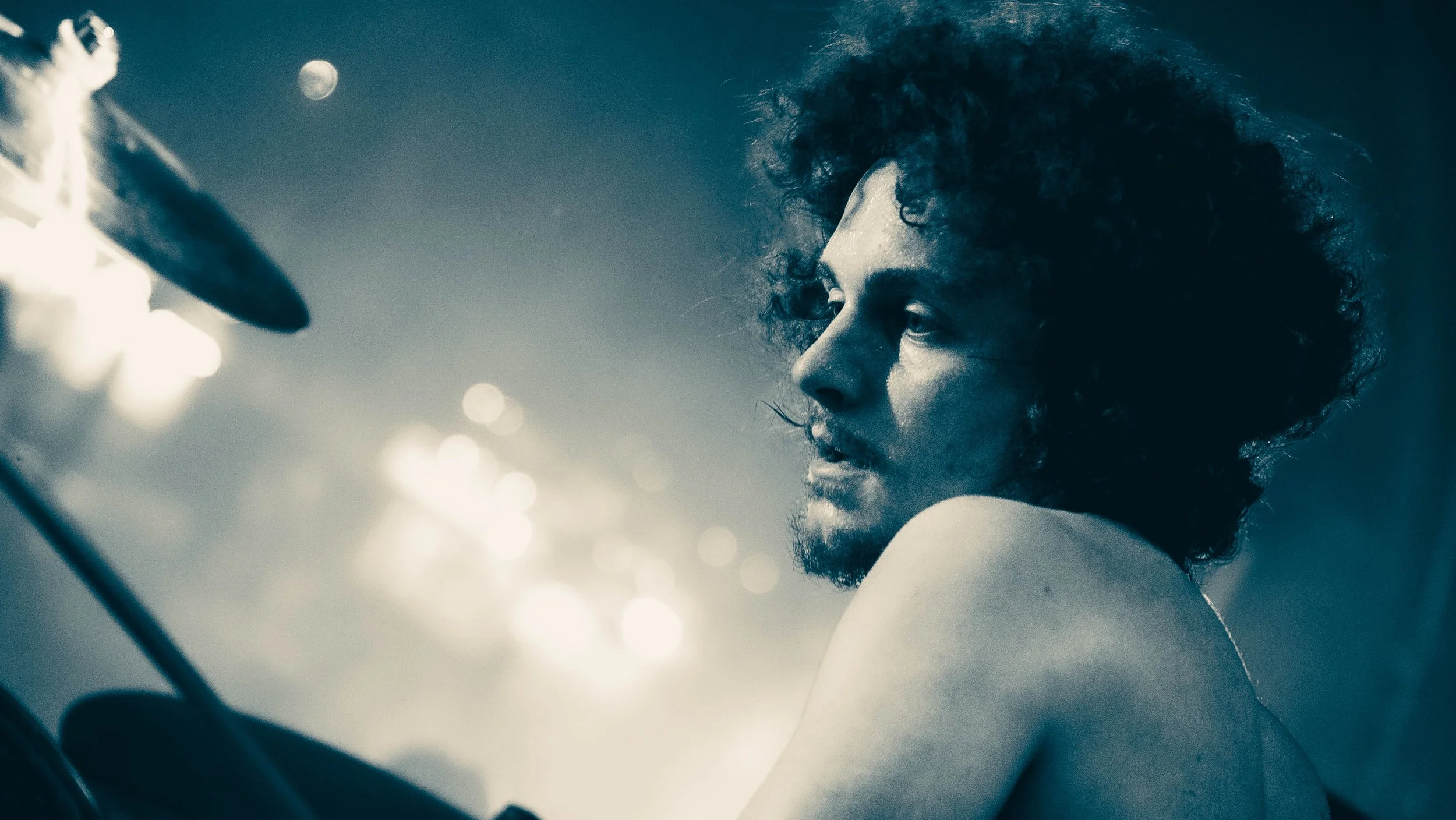 A close-up of a person with curly hair and a beard, looking over their shoulder, with bright lights creating a bokeh effect in the background.