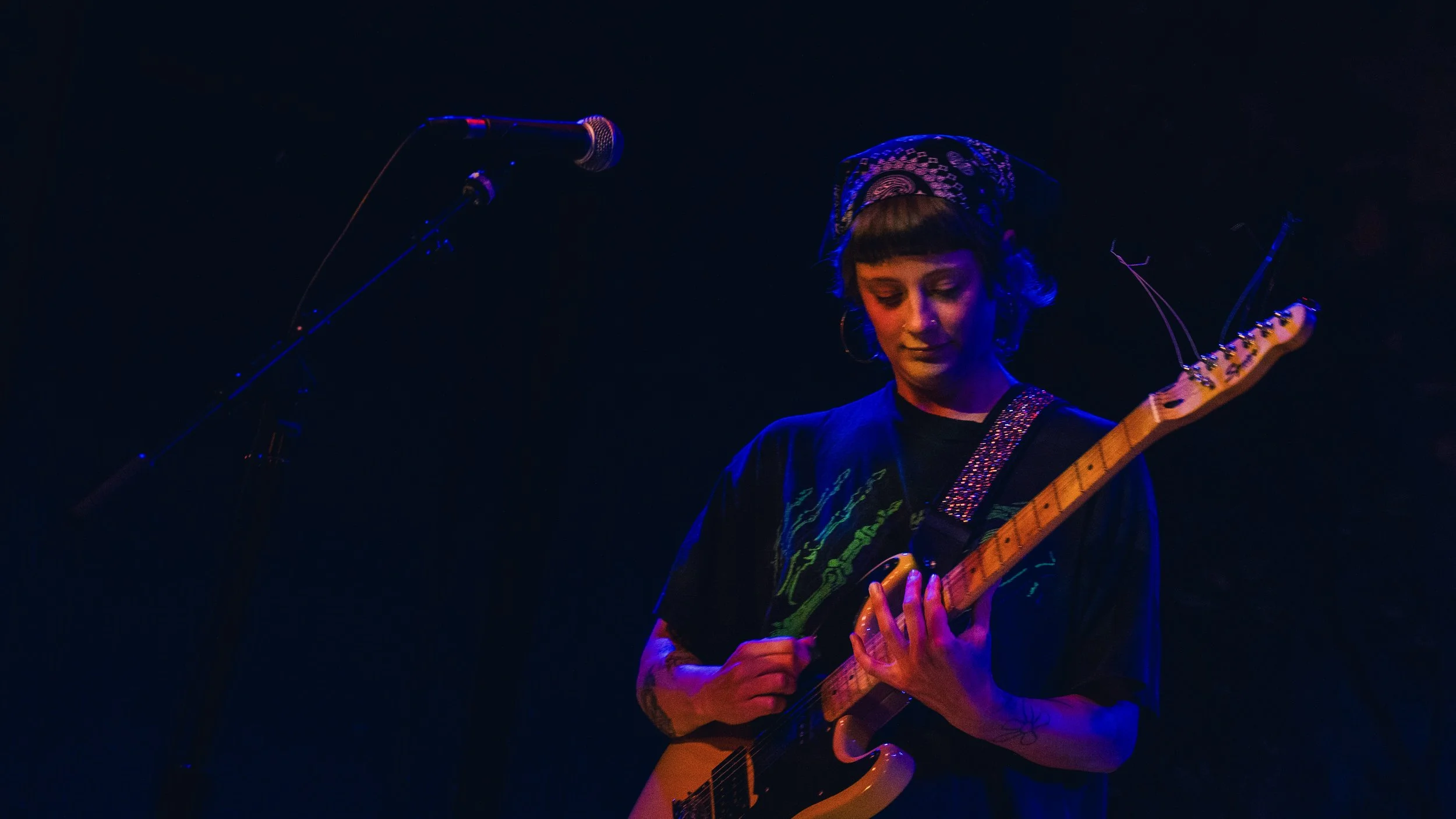 A woman playing an electric guitar on stage under blue and purple lighting, with a microphone nearby.