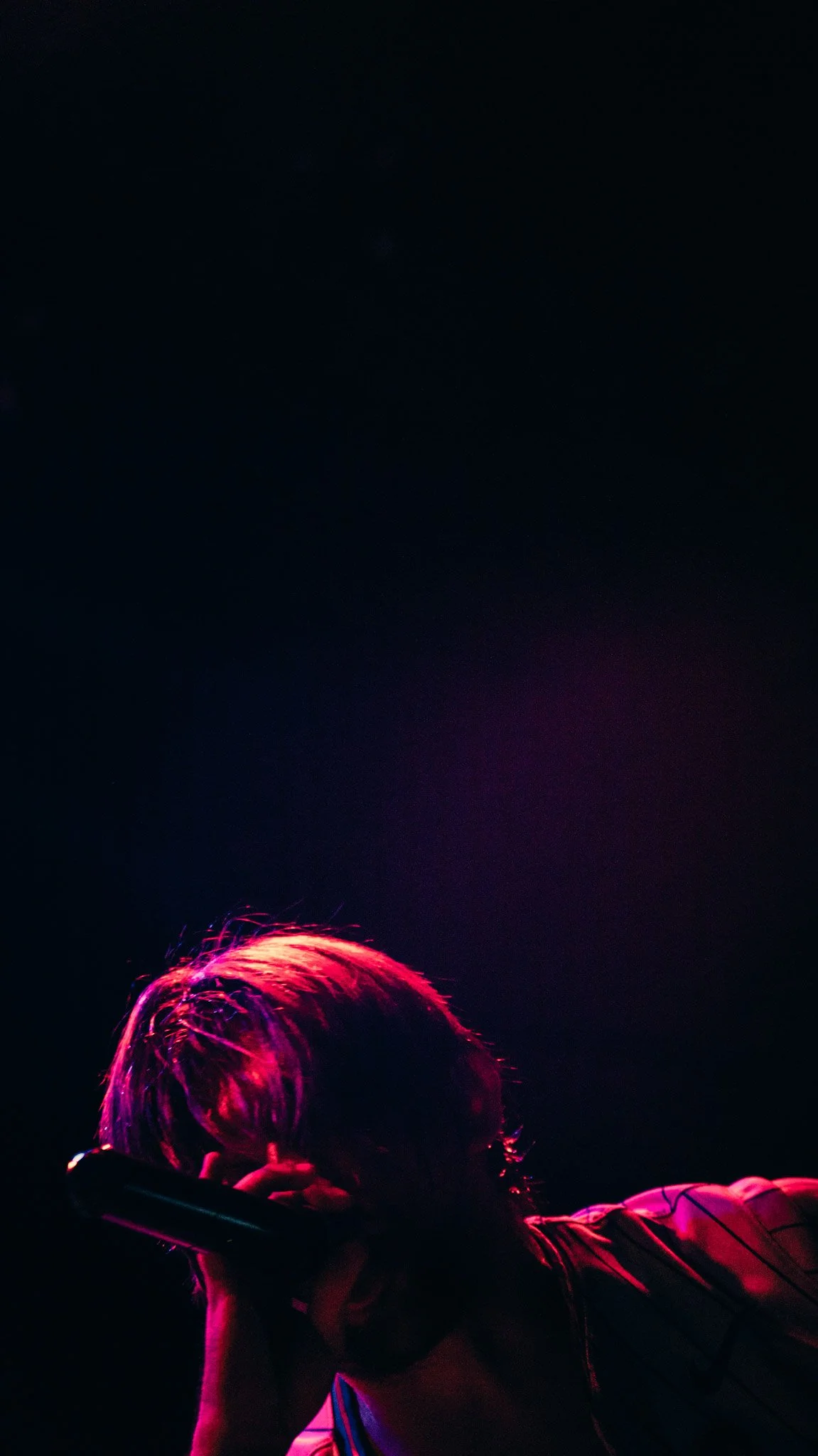 Person with colorful hair holding a microphone in a dark environment with purple and pink lighting.