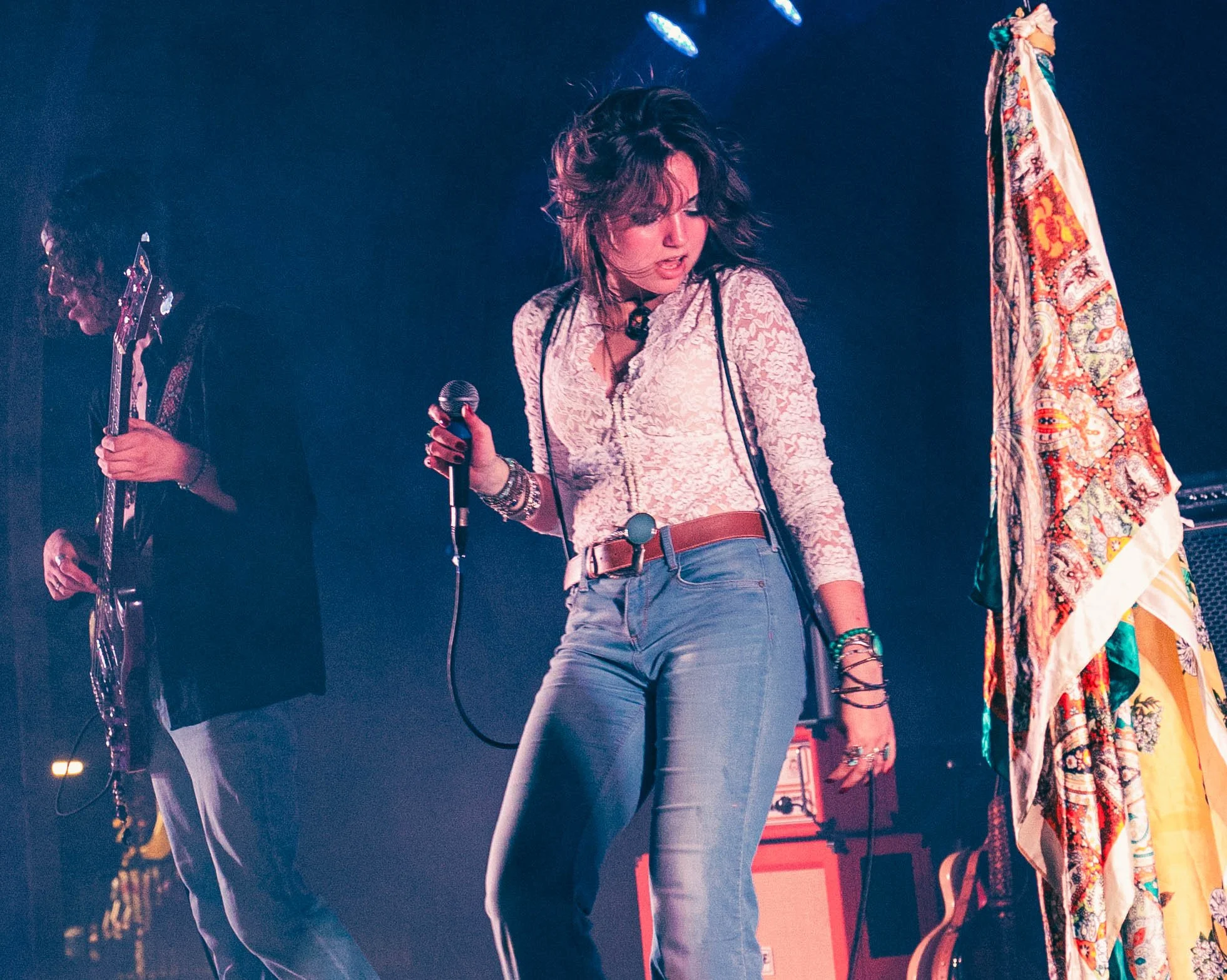A woman singing into a microphone on stage, with a man playing guitar nearby, and colorful stage lighting and fabric in the background.