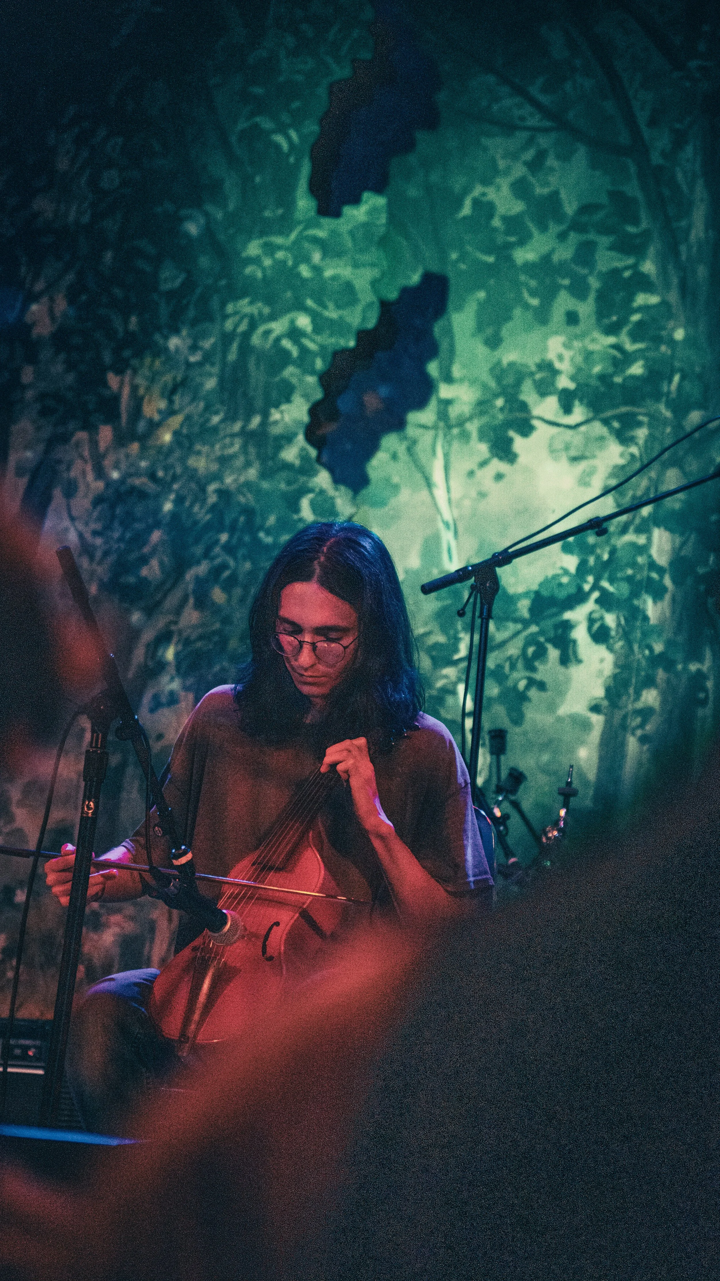 A woman with glasses playing the cello on stage with a colorful, forest-themed backdrop and music stands around her.