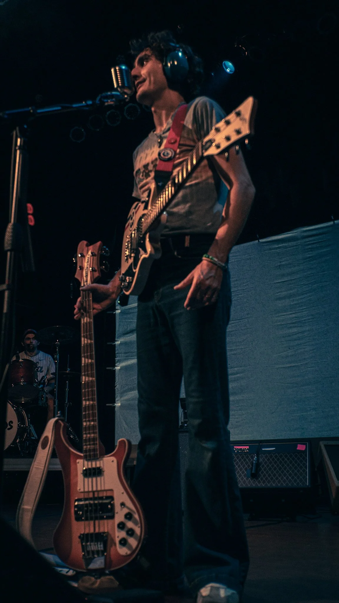 A man wearing headphones is singing into a microphone while playing an electric guitar on stage. A drummer is visible in the background, and there are additional guitars and equipment on stage.