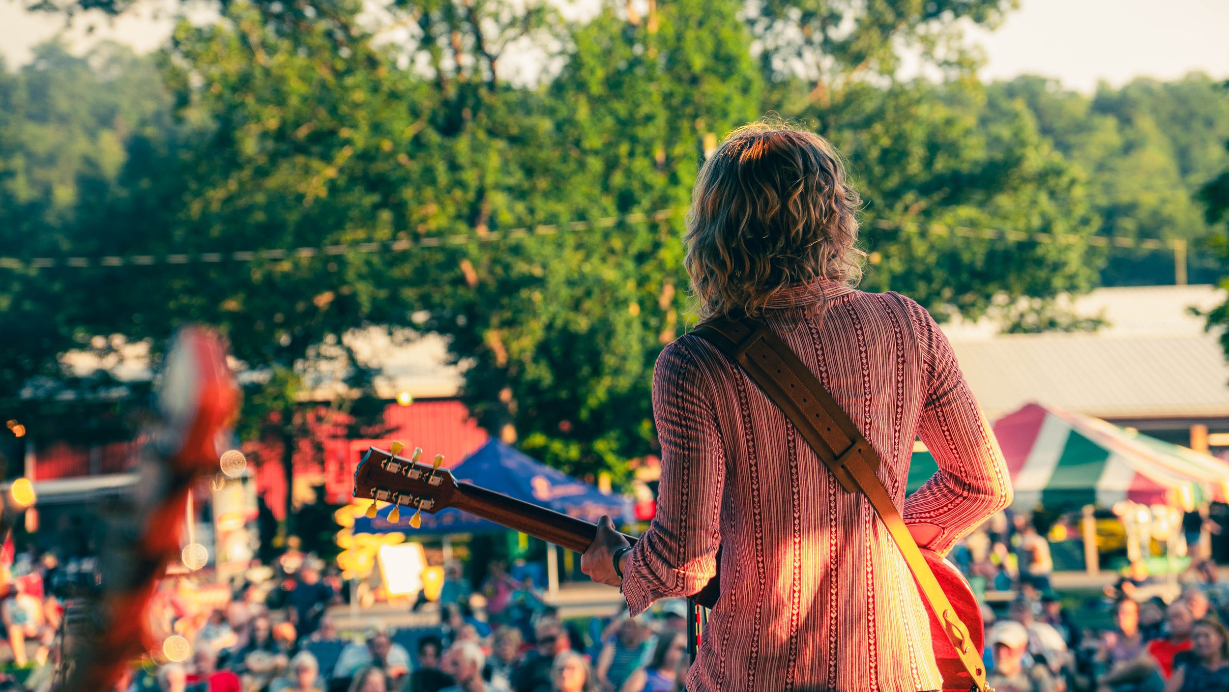 Person with curly hair wearing a striped shirt playing guitar at an outdoor event with trees and colorful tents in background.