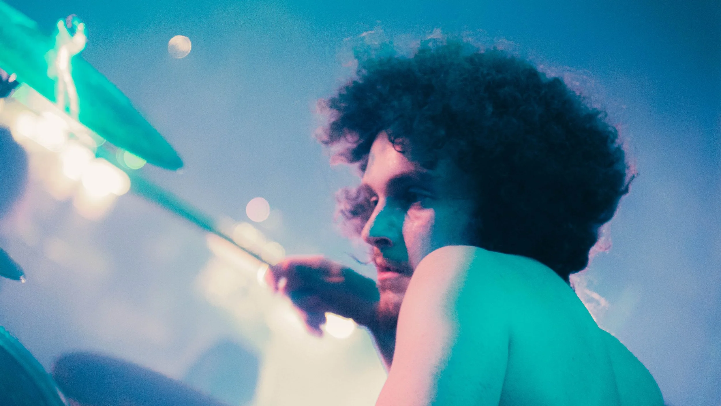 A young woman with curly hair looks at her phone, with a blurred cityscape and moon in the background, illuminated by colorful neon lights.