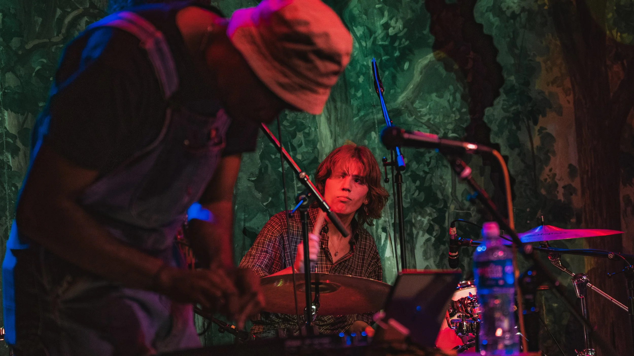 Two musicians performing on stage with a green leafy background. One standing with a hat and the other sitting with shoulder-length hair, playing drums. Stage lighting casts colorful highlights.