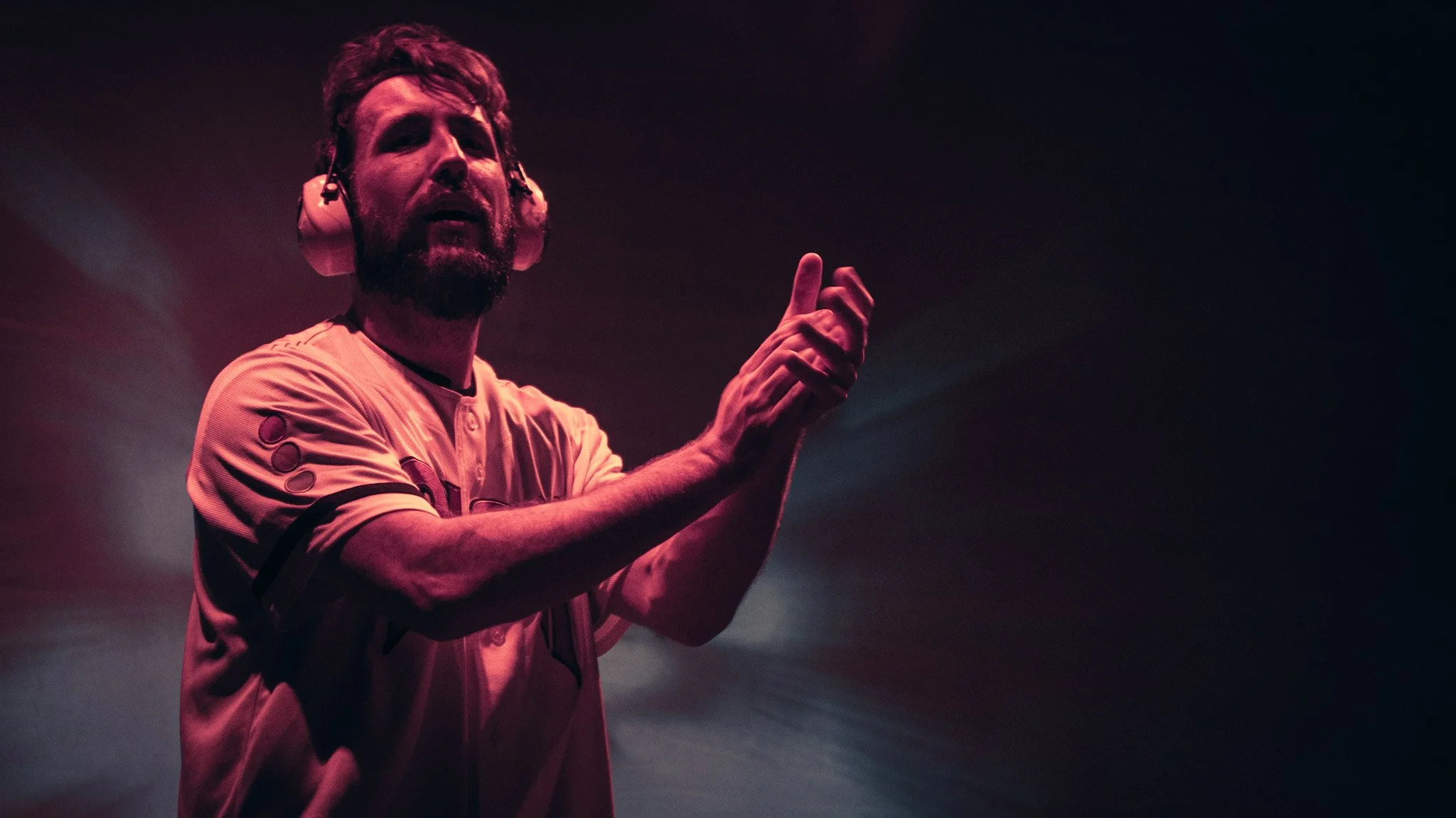 A man with a beard wearing headphones and a sports jersey, gesturing with his hands while performing on stage with dark background.