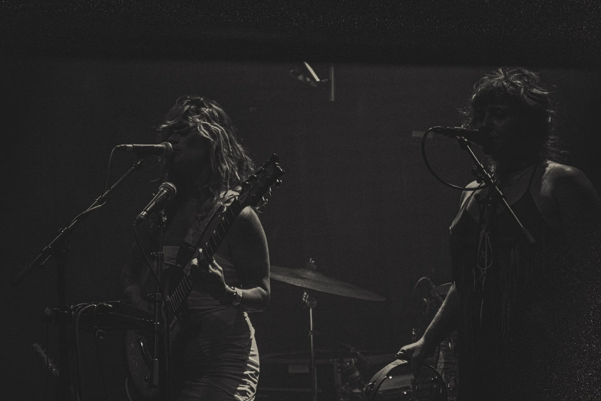 Two women performing on stage in a dark setting, one playing guitar and the other singing into a microphone.