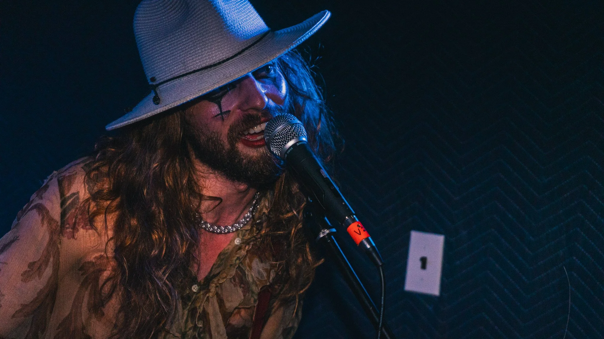 A man with long hair and a beard wearing a wide-brimmed hat, glasses, and a chain necklace, singing into a microphone in a dimly lit room with black soundproof walls.