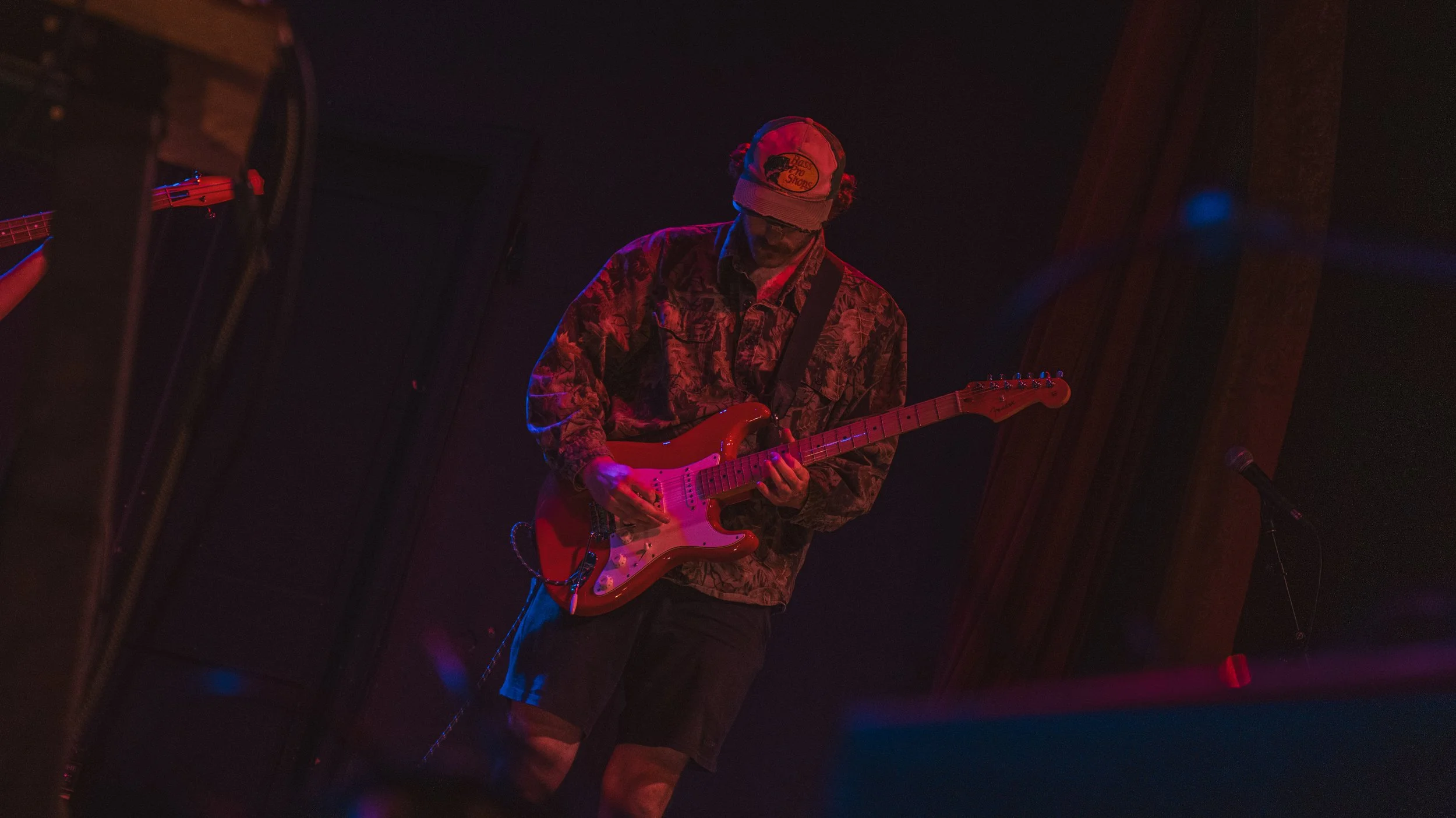 A person wearing a NASA cap and patterned jacket playing an electric guitar in a dimly lit setting with red and blue stage lighting.