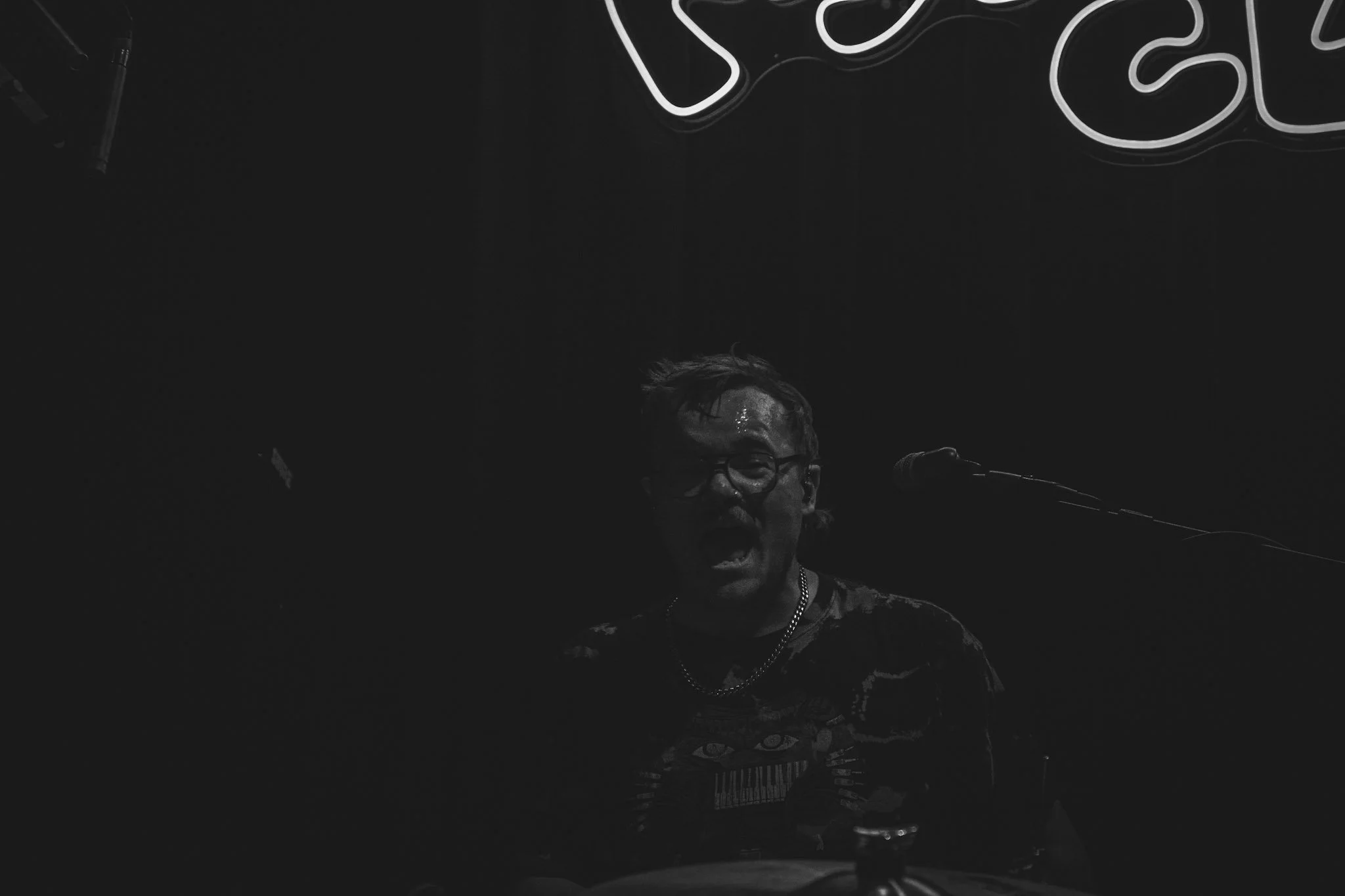 A person with glasses singing or speaking into a microphone on stage, illuminated by neon lights that spell out 'YES' against a dark background.