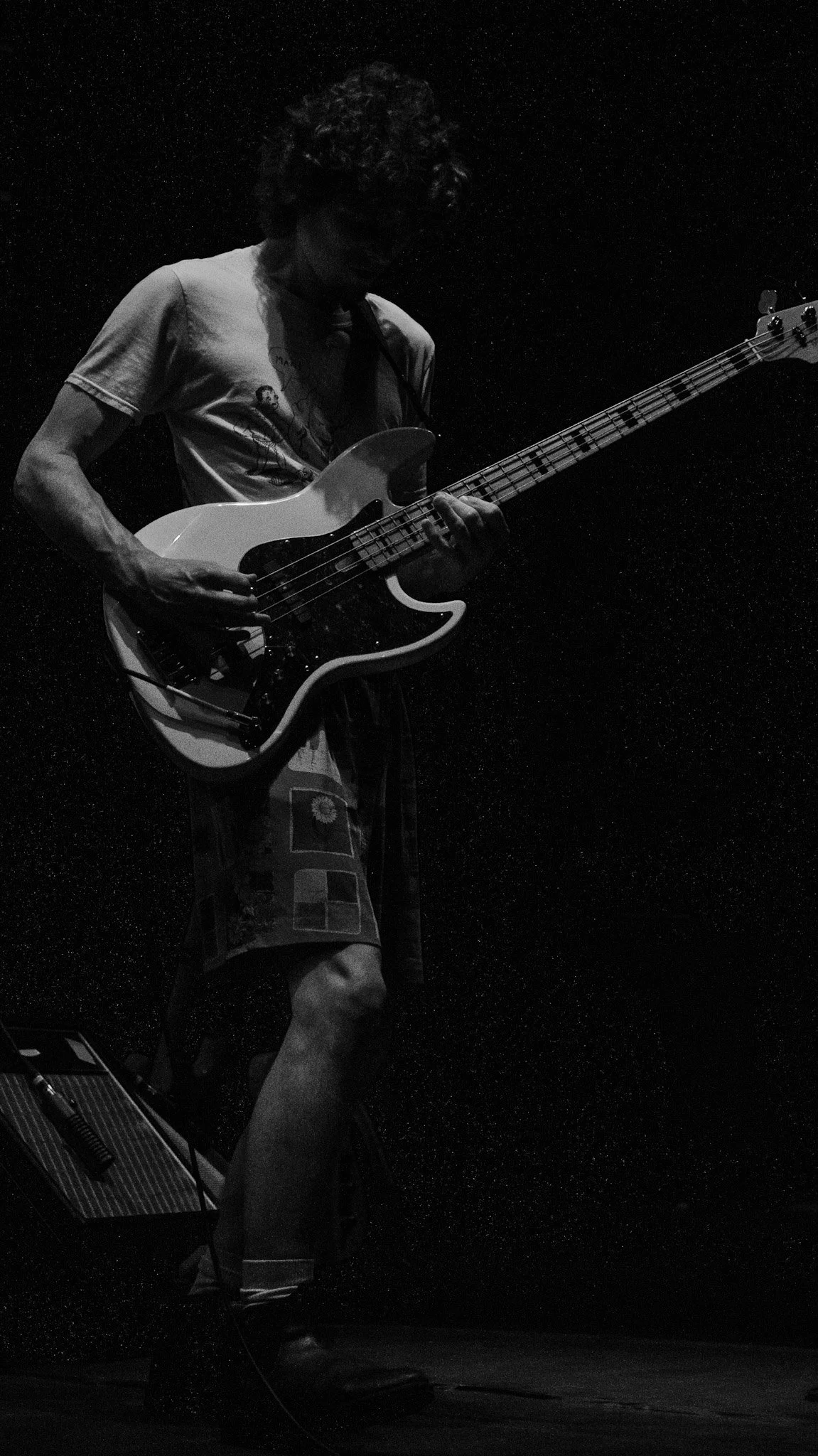 A person playing an electric bass guitar on stage in a dark setting, wearing a T-shirt and patterned shorts.