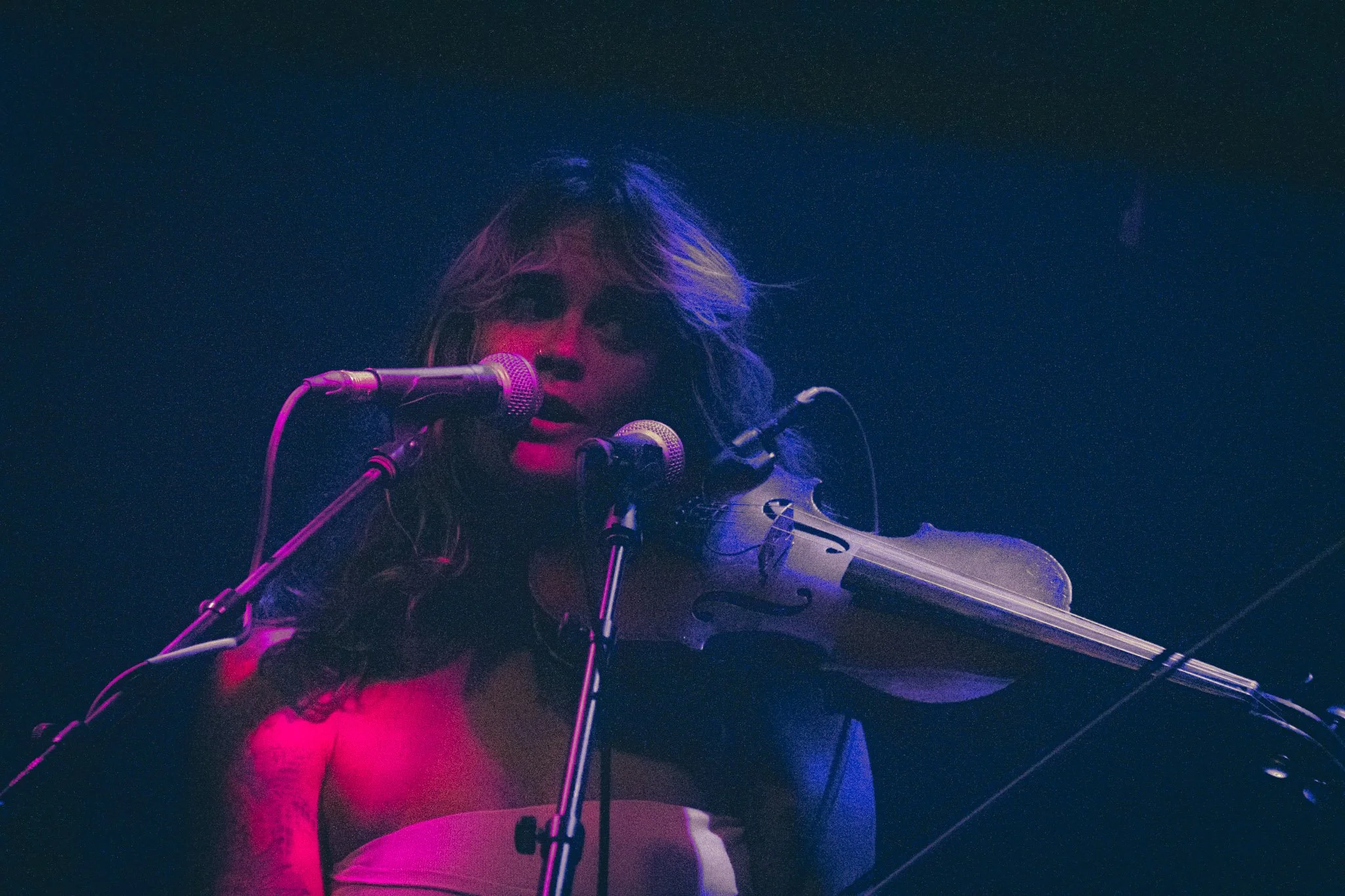 A woman with long hair playing a violin and singing into a microphone on stage, illuminated by vibrant multicolored stage lighting.
