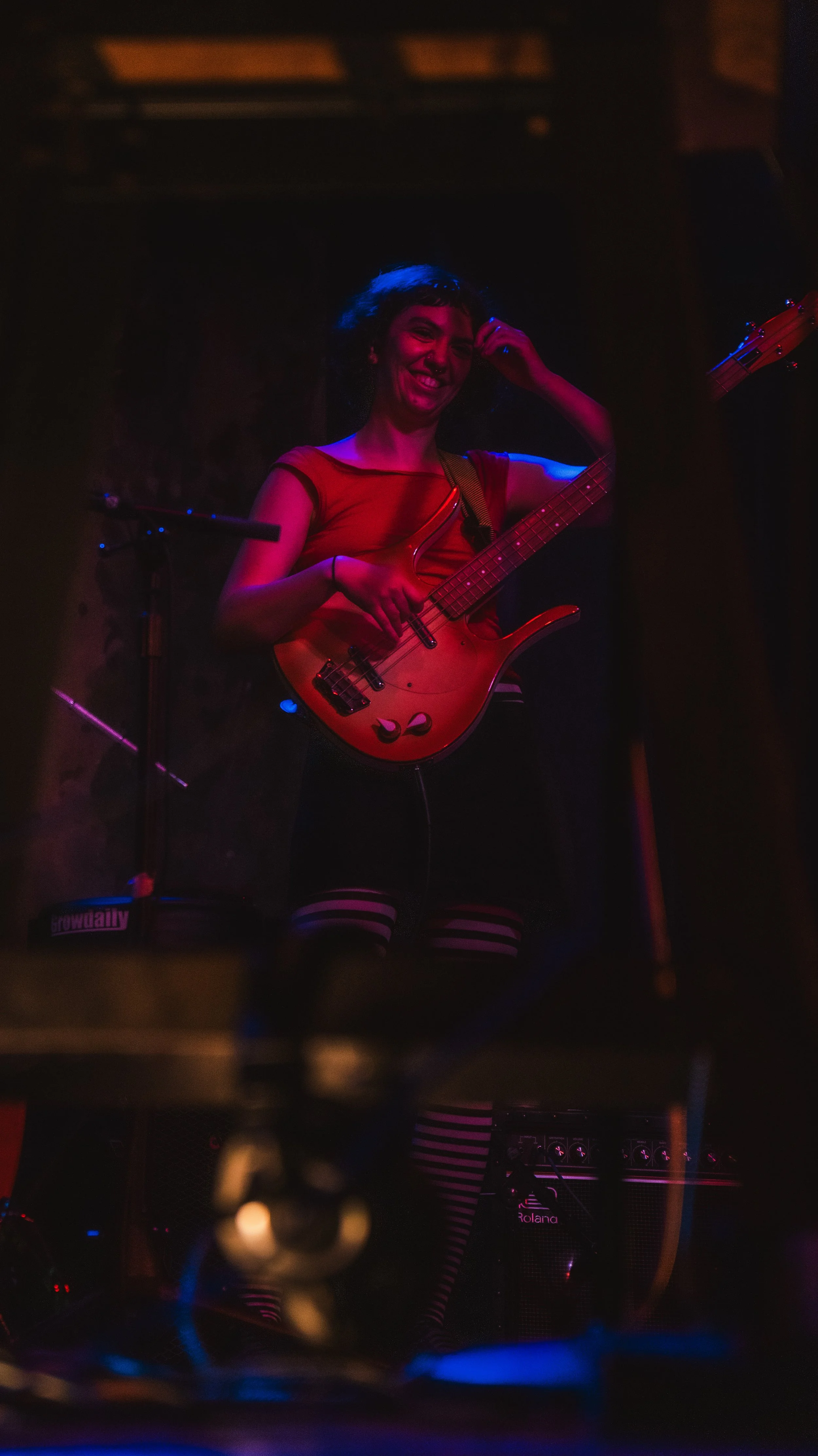 A woman playing an electric bass guitar on stage, smiling, wearing a red top and striped thigh-high socks, illuminated by colorful stage lighting.