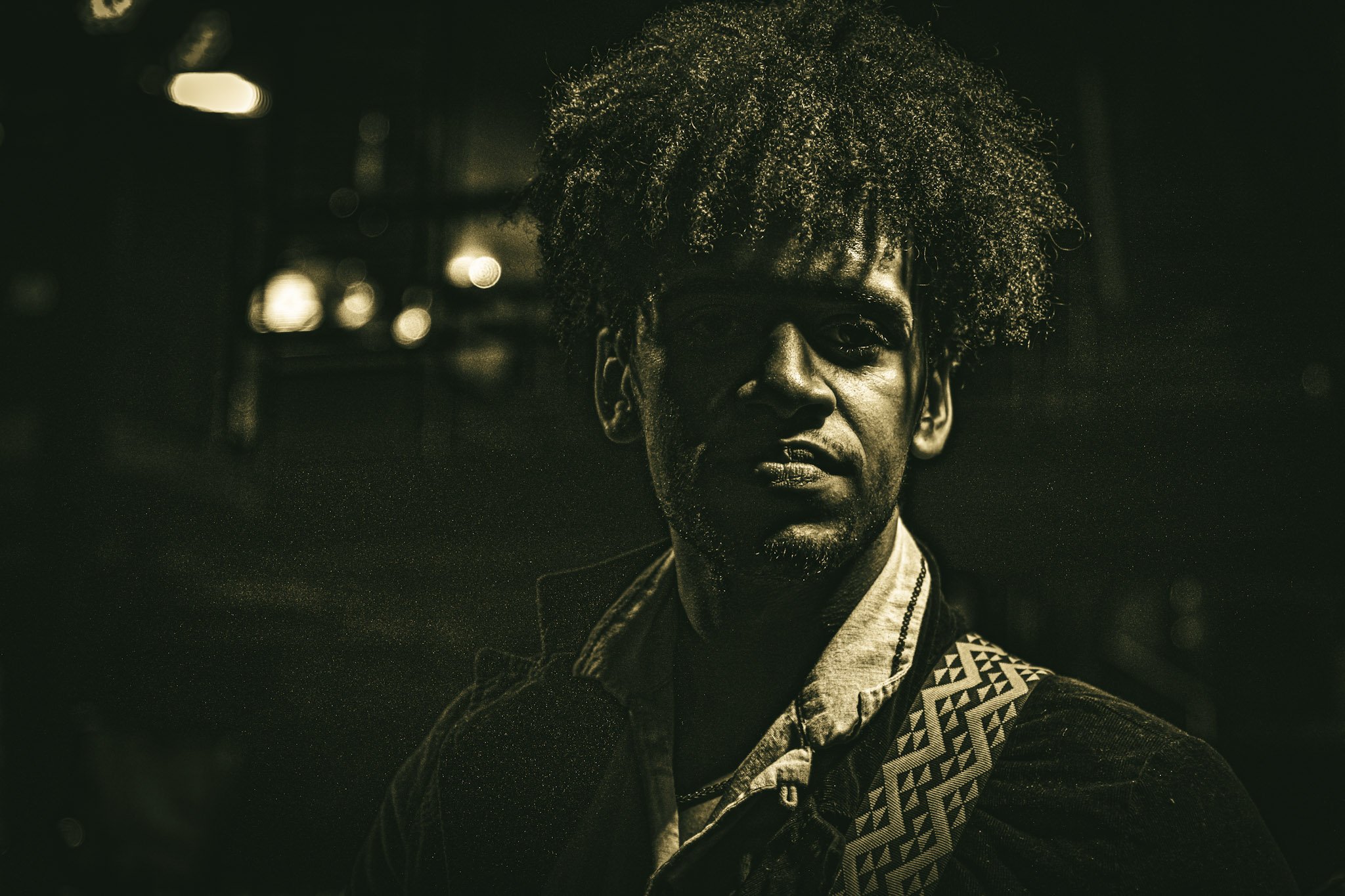 A portrait of a young man with dark curly hair, wearing a patterned jacket and a light shirt, standing outdoors at night with blurred city lights in the background.