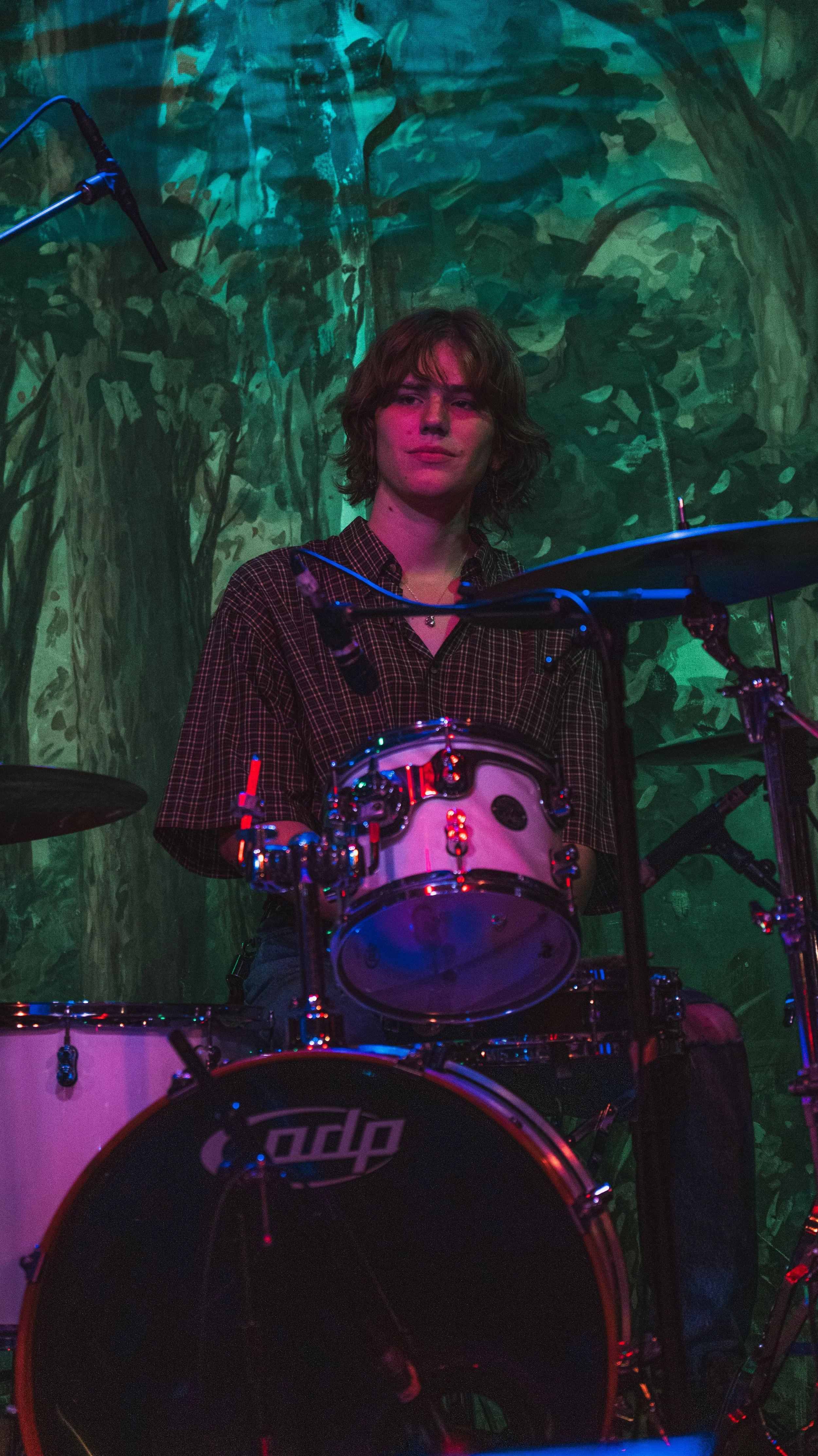 A person with shoulder-length brown hair sitting behind a drum set in a dimly lit environment with green and pink lighting, and a painted or projected background resembling a forest scene.