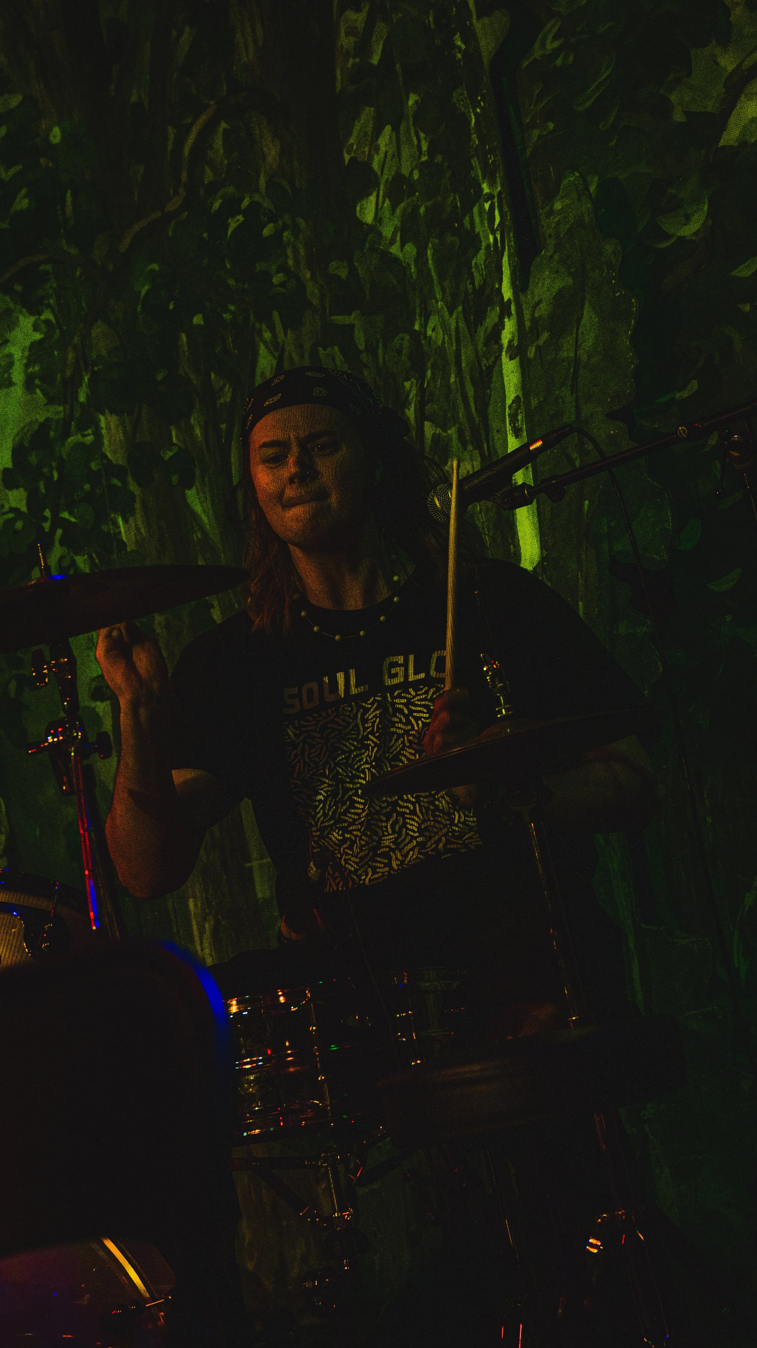 A female drummer performing on stage with green lighting and a patterned backdrop.