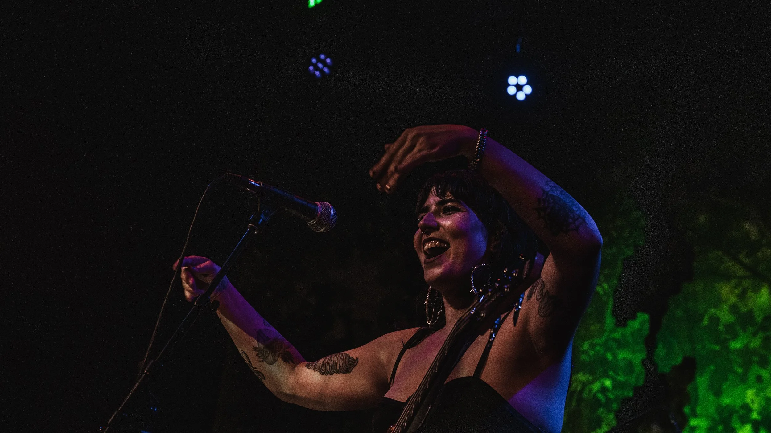A woman with tattoos on her arms singing or speaking into a microphone on stage, with her arms raised, illuminated by stage lights, dark background with some green lighting.
