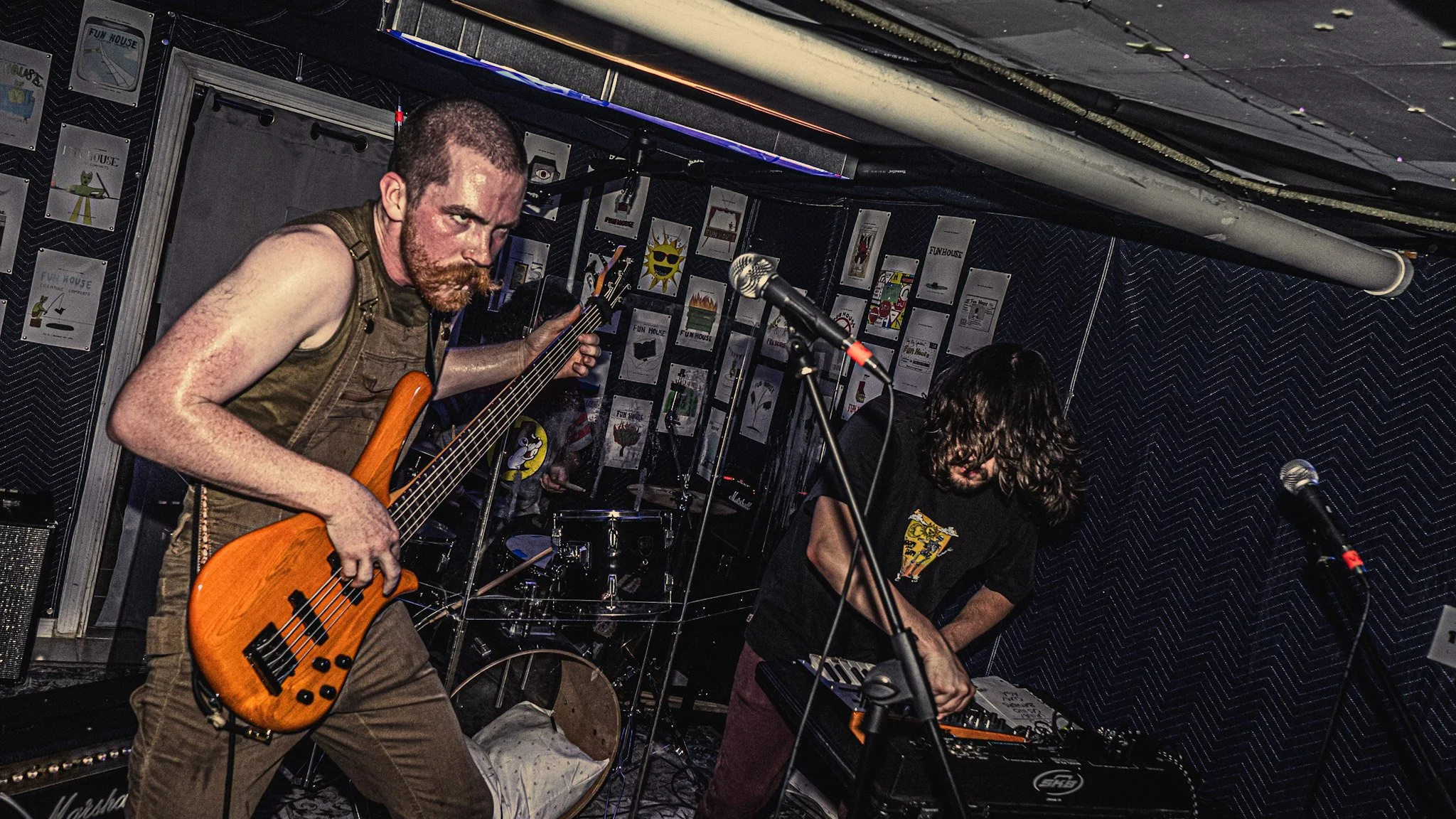 Two musicians performing in a small, dimly lit venue. The man on the left is playing an orange bass guitar, with a focused expression and sweaty face, wearing a sleeveless shirt. The man on the right is deeply engrossed in playing a keyboard, with lo
