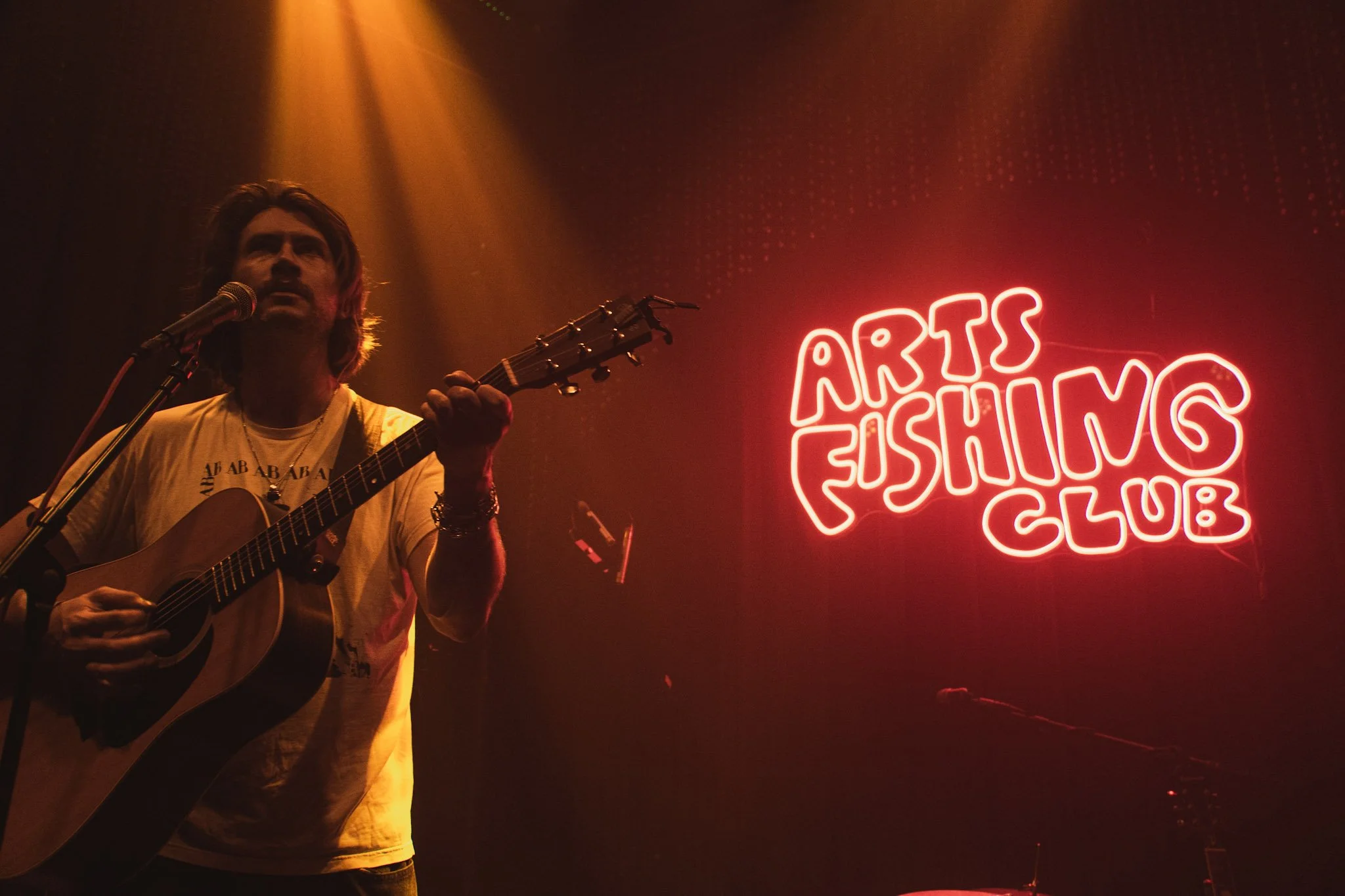 A man playing an acoustic guitar and singing into a microphone on stage, illuminated by warm stage lights, with a bright red neon sign behind him that reads 'ARTS FISHING CLUB'.
