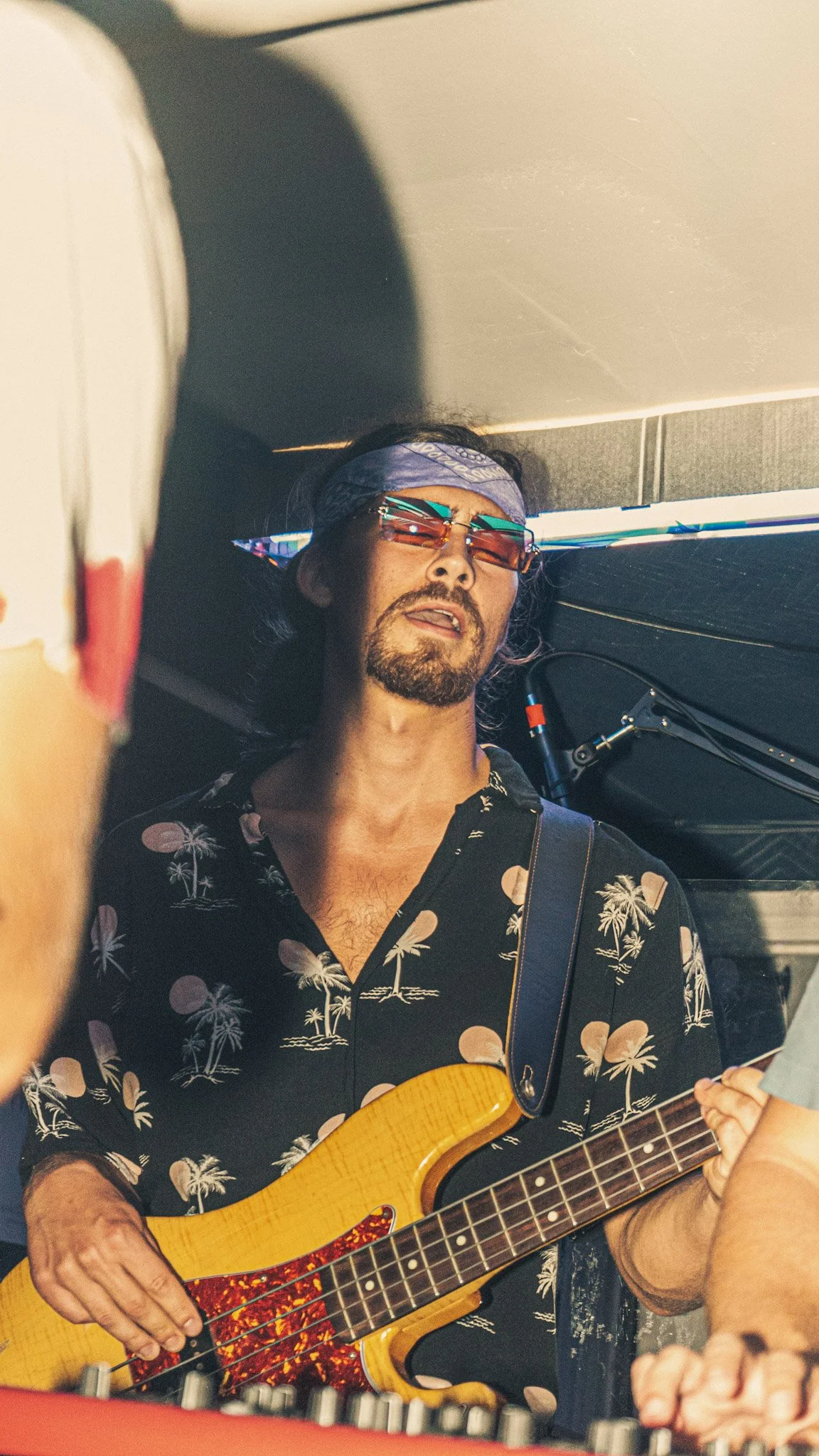 A man with dark hair, sunglasses, a patterned shirt, and a bandana on his head is playing a yellow acoustic guitar, with a person nearby playing an instrument or adjusting equipment.
